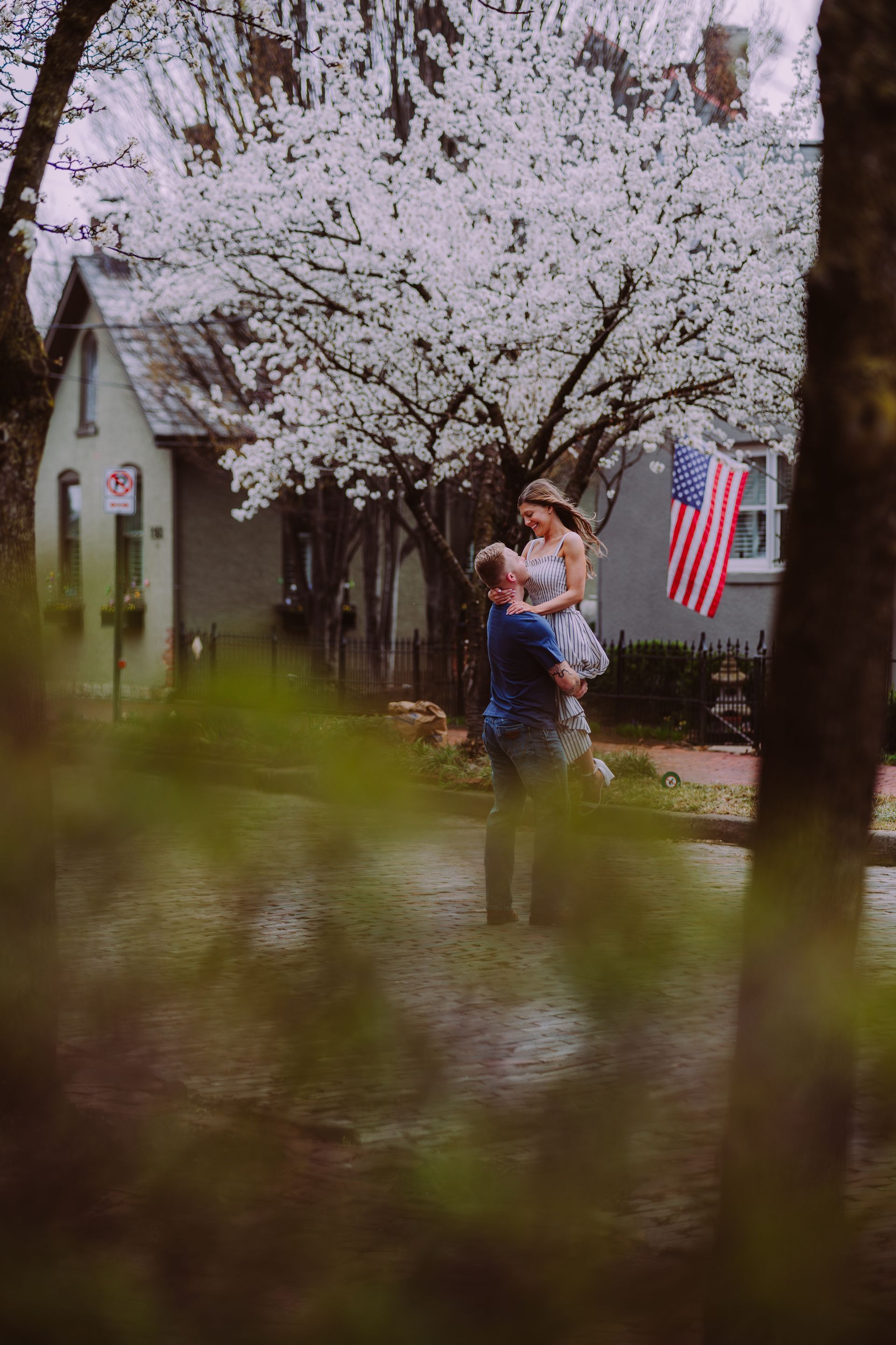 A couple embraces, with one person lifting the other, standing on a street in front of a blooming tree and a house.