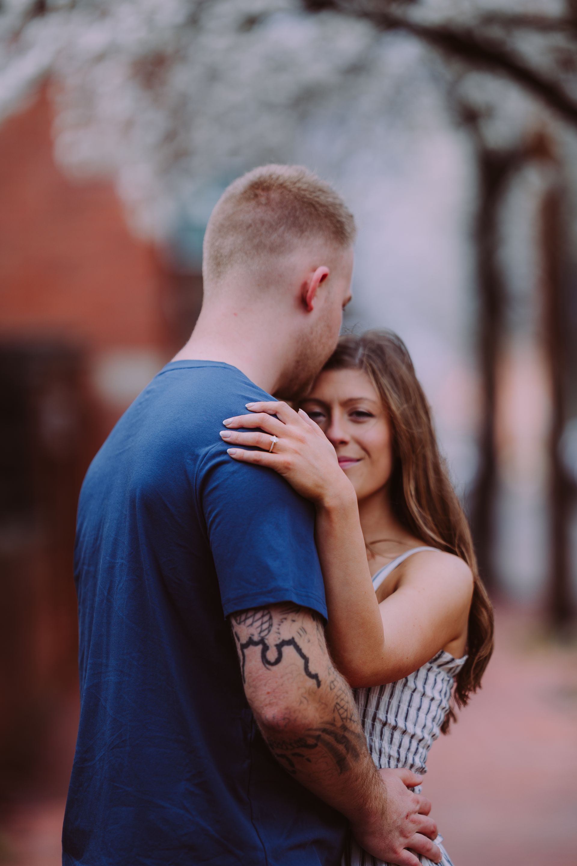 A person in a blue t-shirt hugs someone in a patterned sundress outdoors, with a blurred, sunlit background.