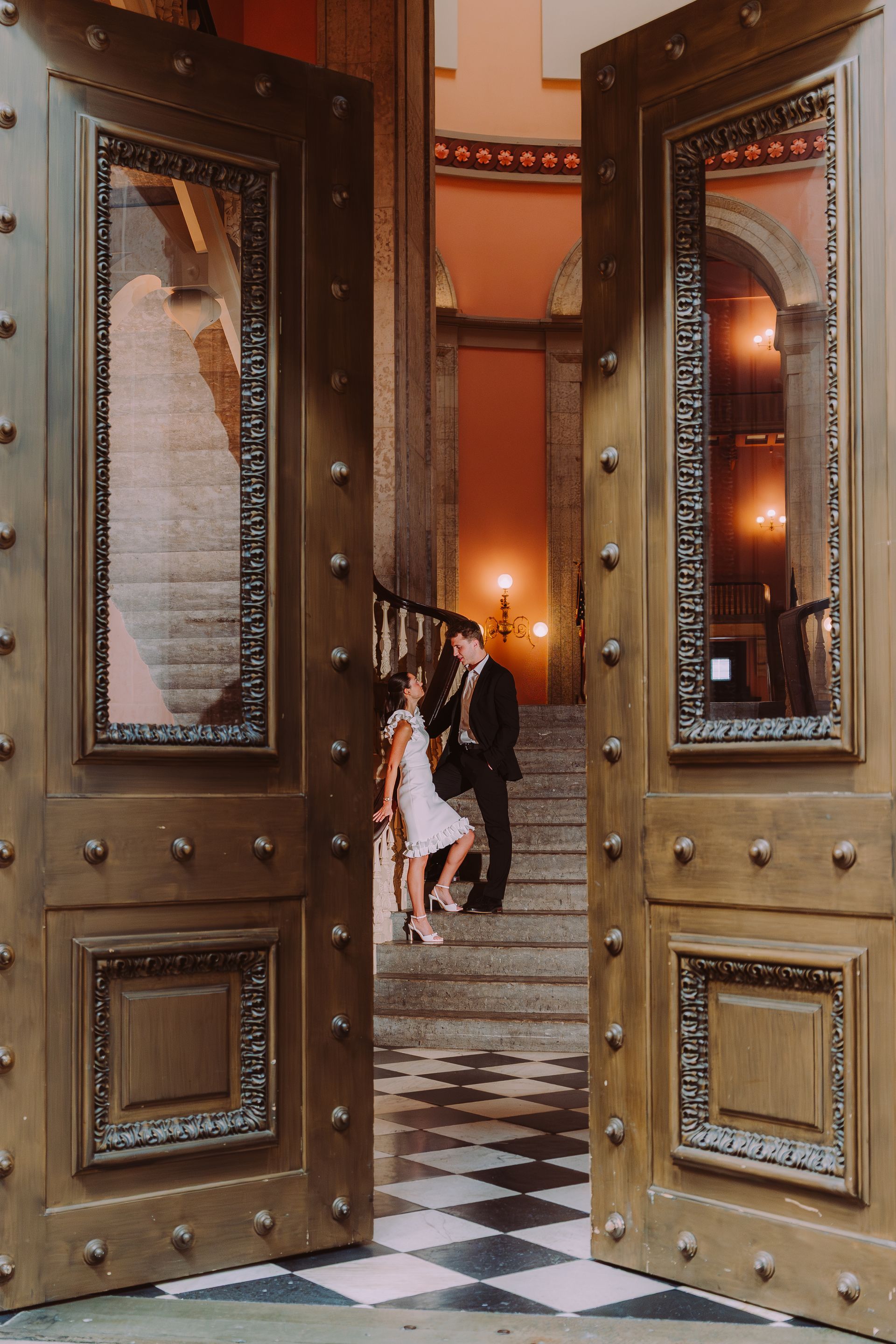 A couple poses on stone stairs inside a grand building, viewed through a large, open set of ornate metal doors.