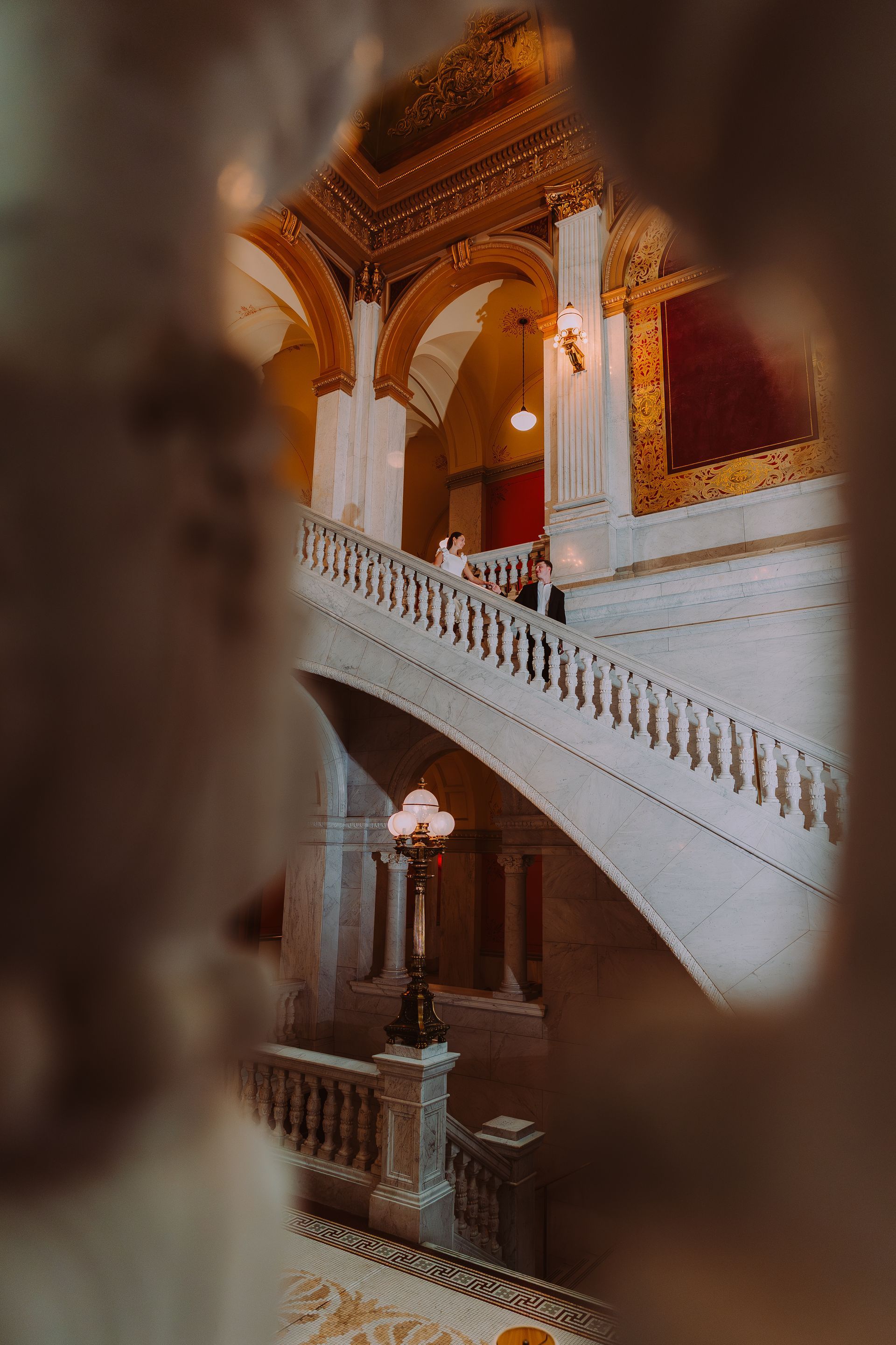 A grand, ornate staircase with white marble railings is framed by a blurred foreground in a historic building.