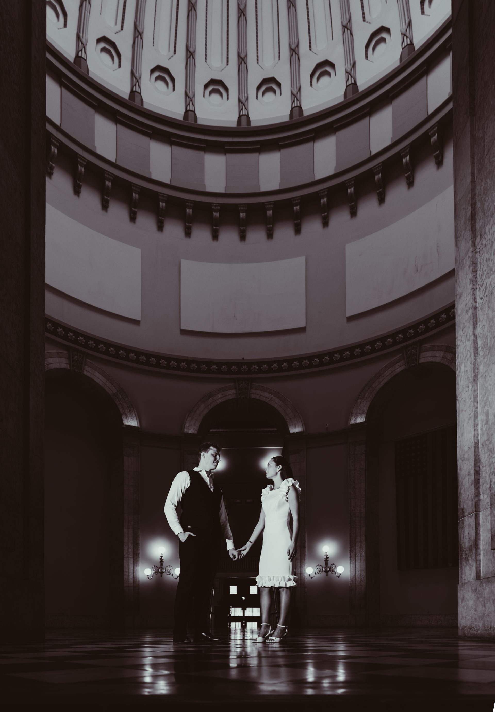 A couple holds hands standing in the center of a grand, dimly lit domed building with classic architectural details.