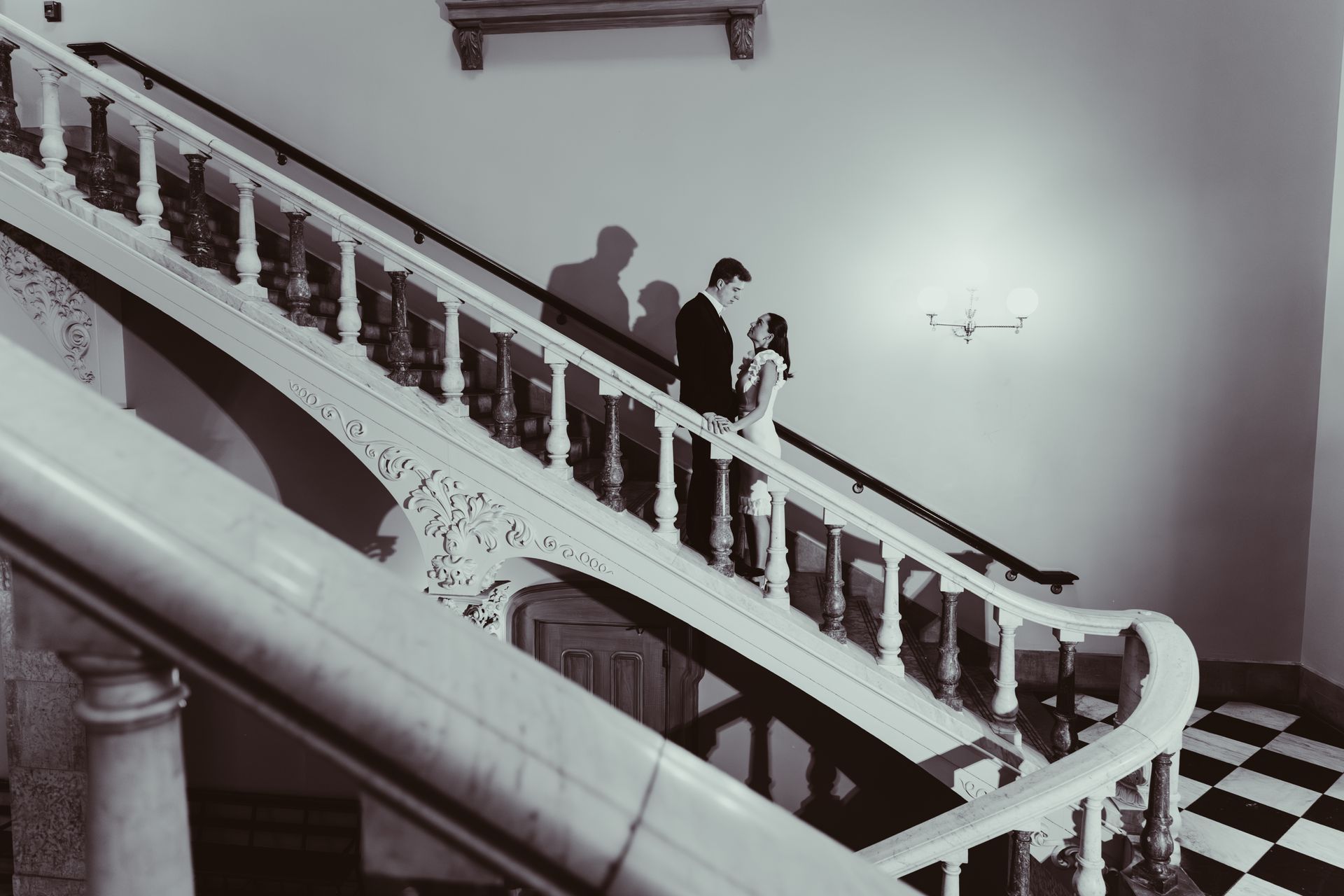 A couple stands on an elegant, curved staircase with a checkered floor, casting shadows on the wall behind them.