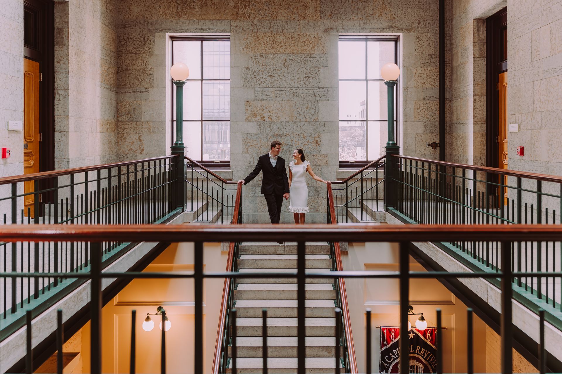 A couple stands together on a stone staircase inside a grand building, framed by symmetric windows and railings.