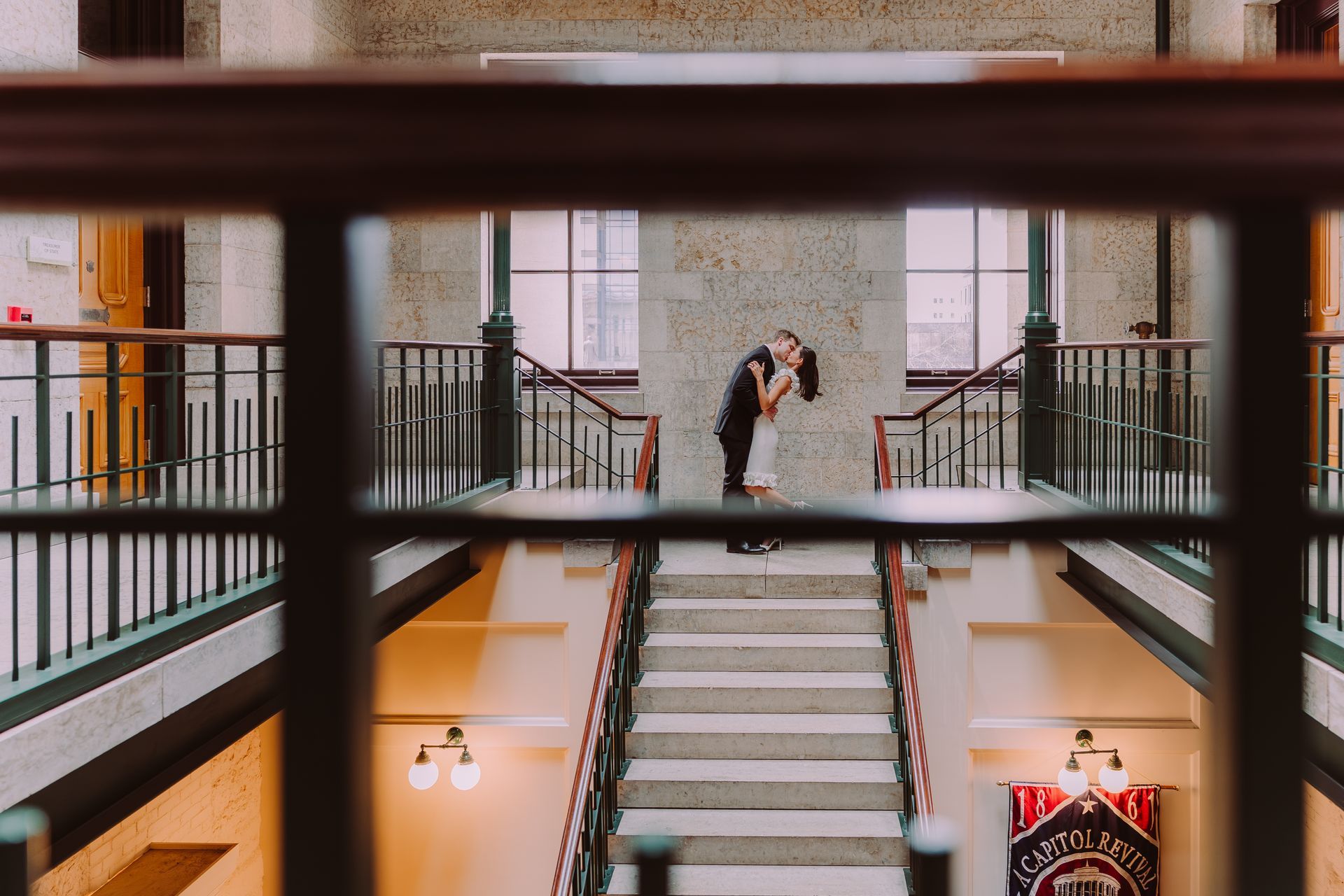 A couple kisses on a staircase landing, viewed through a foreground railing in a building with stone walls and wood rails.