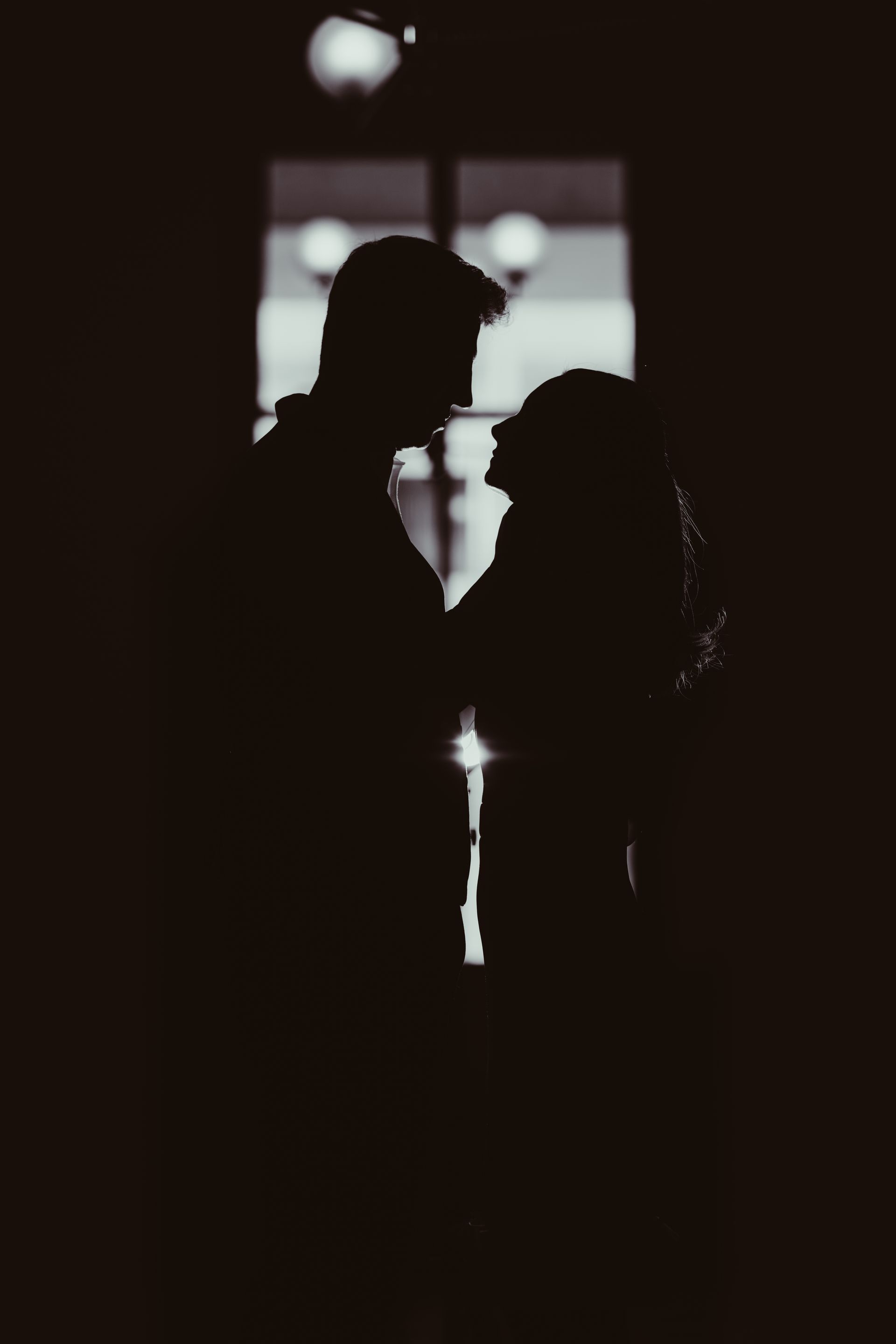 A silhouette of a couple standing face-to-face against a bright, multi-paned window.
