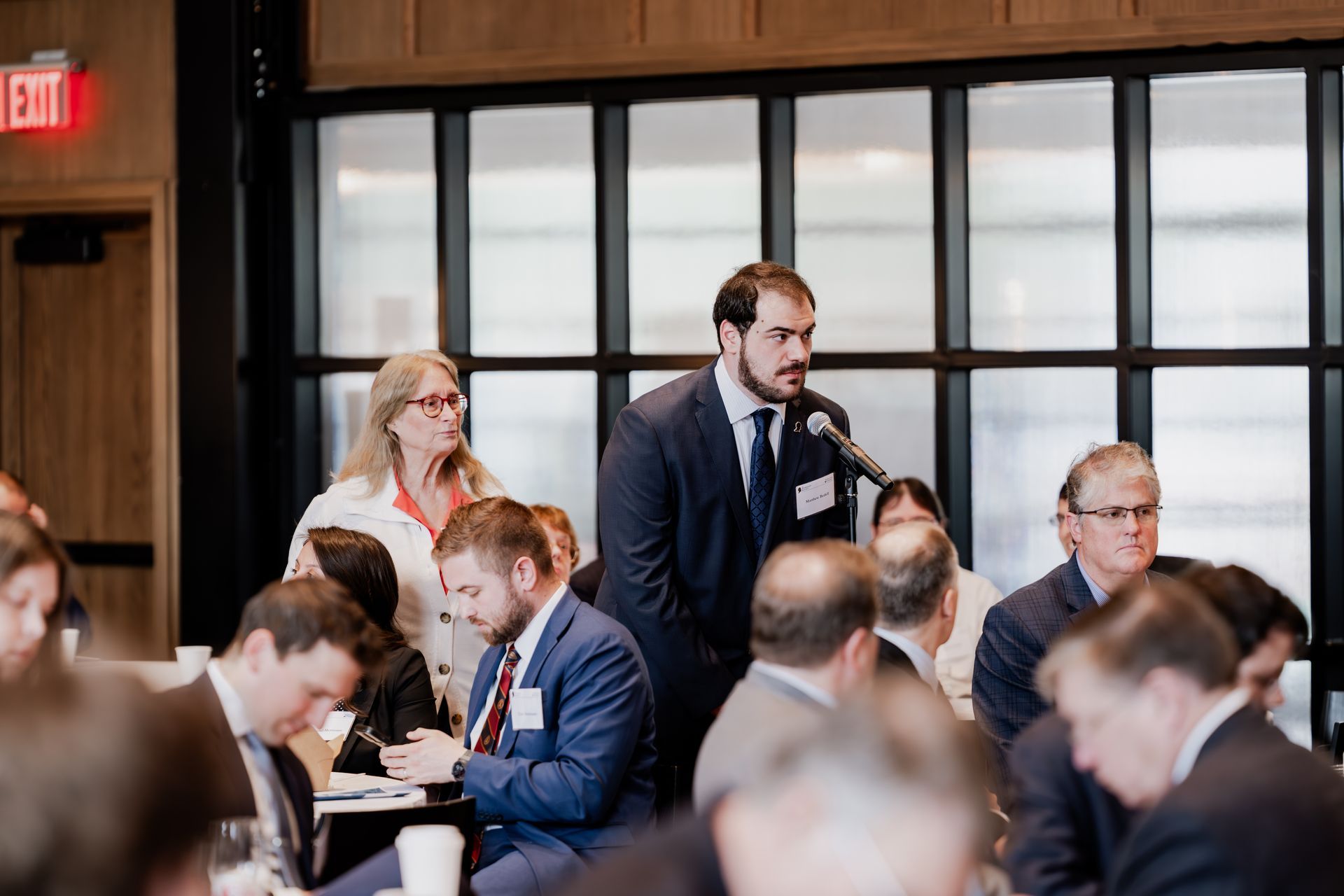 A professional in a suit speaks into a microphone at a meeting while others listen in a room with window-pane dividers.