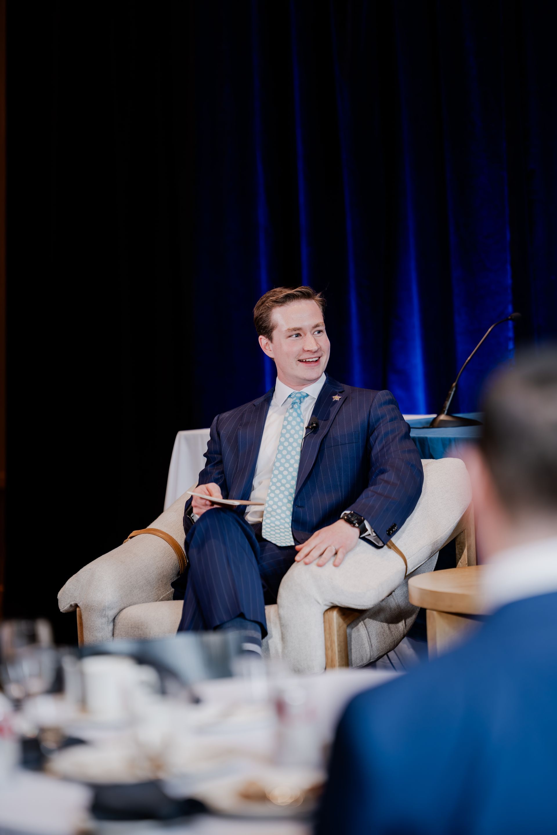 A professional in a suit and light blue tie speaks while seated in an armchair on stage against a dark blue backdrop.