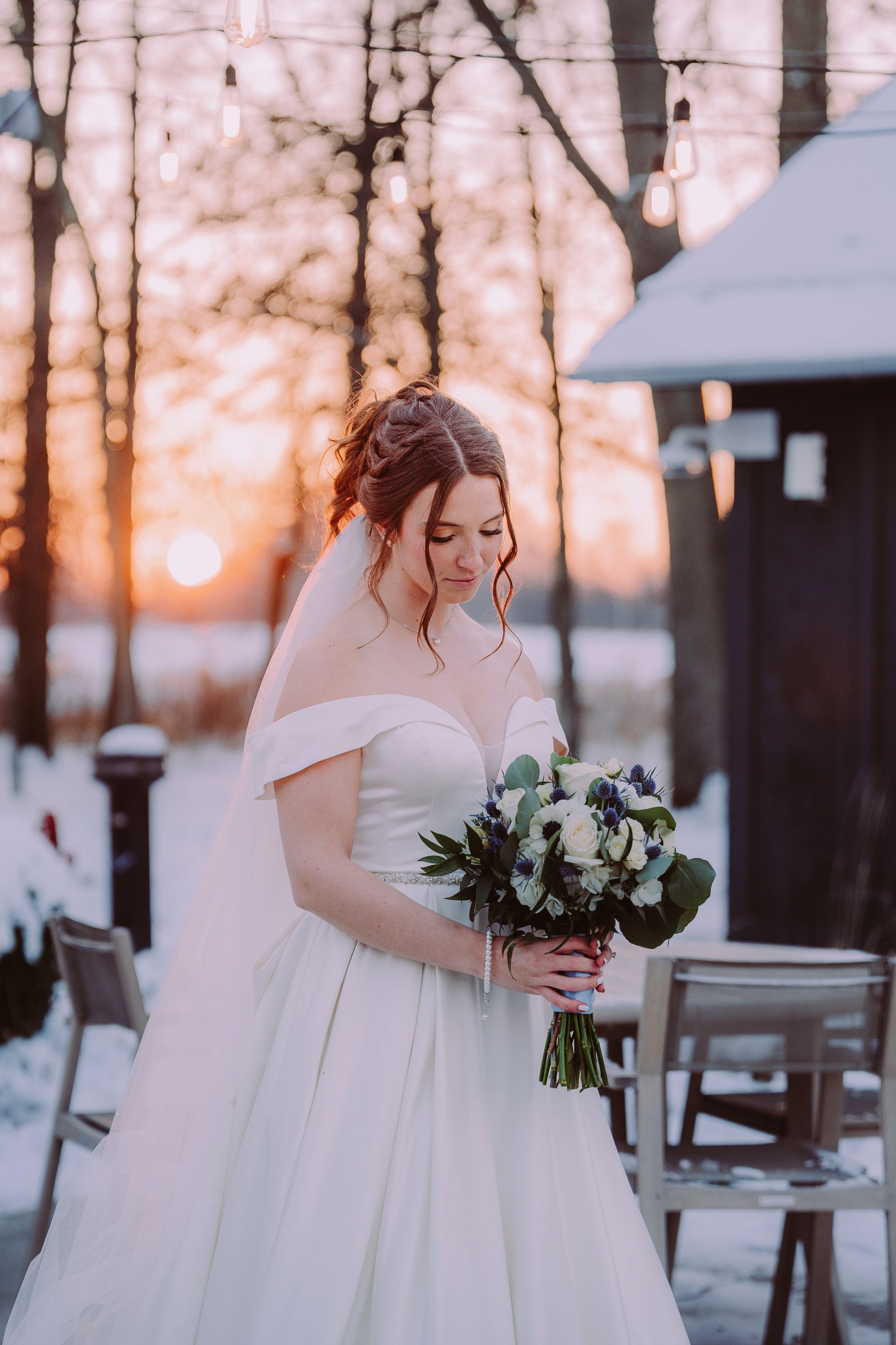 Bride in white dress holding flowers, snowy outdoor setting at sunset.