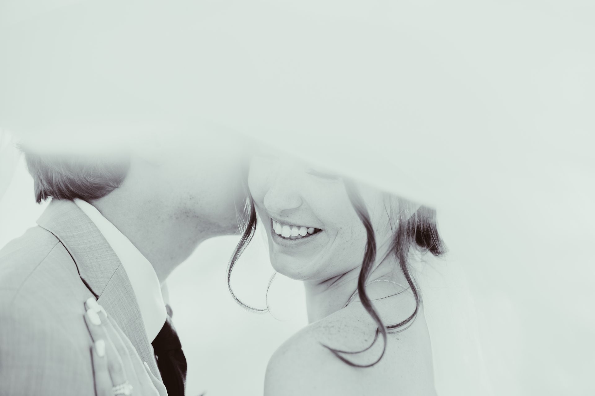 Man kissing a smiling person on the cheek; they are partially obscured by a veil, suggesting a wedding.