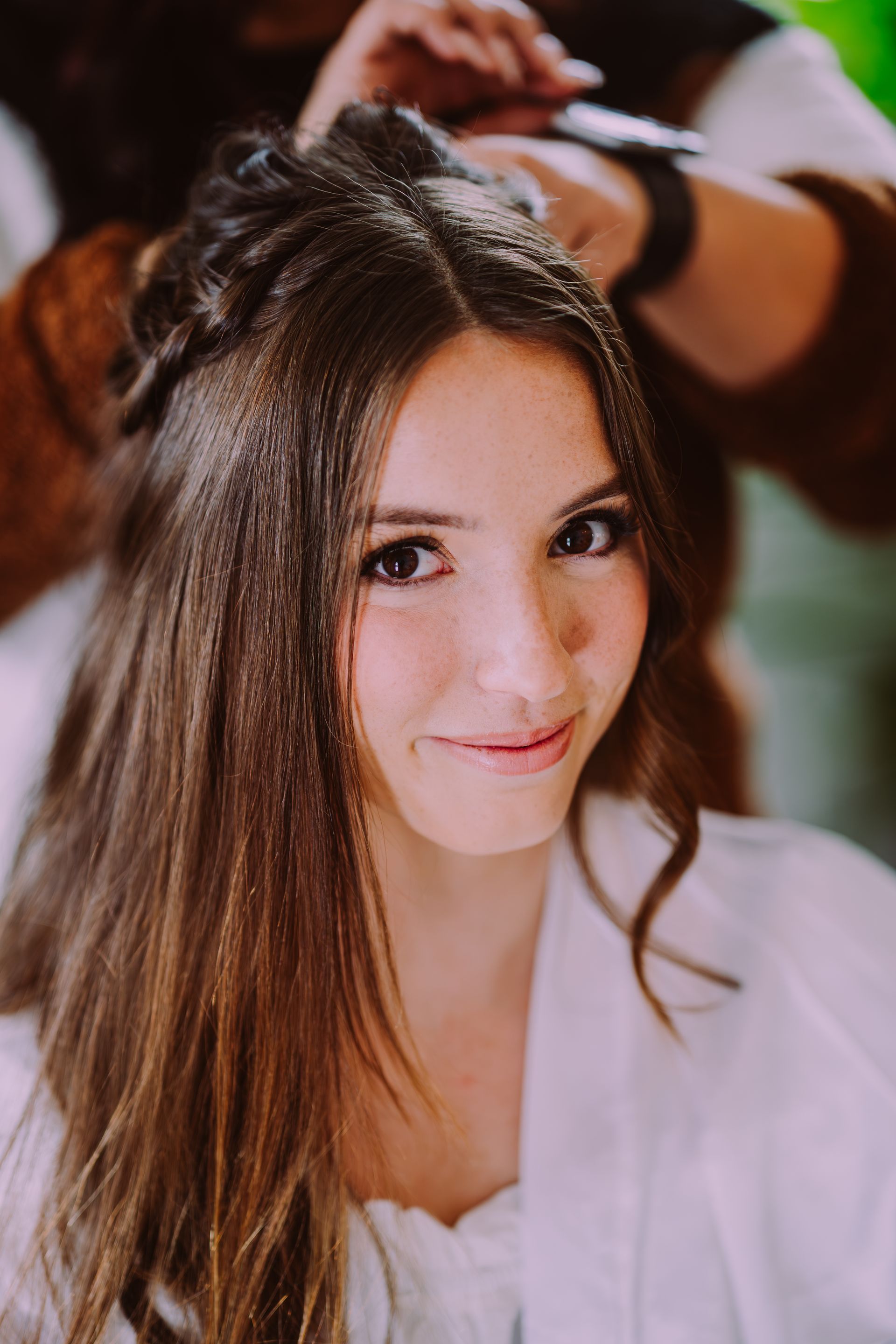 Woman with long brown hair, smiling, having hair styled. Person's hands, watch, and coat visible in the background.