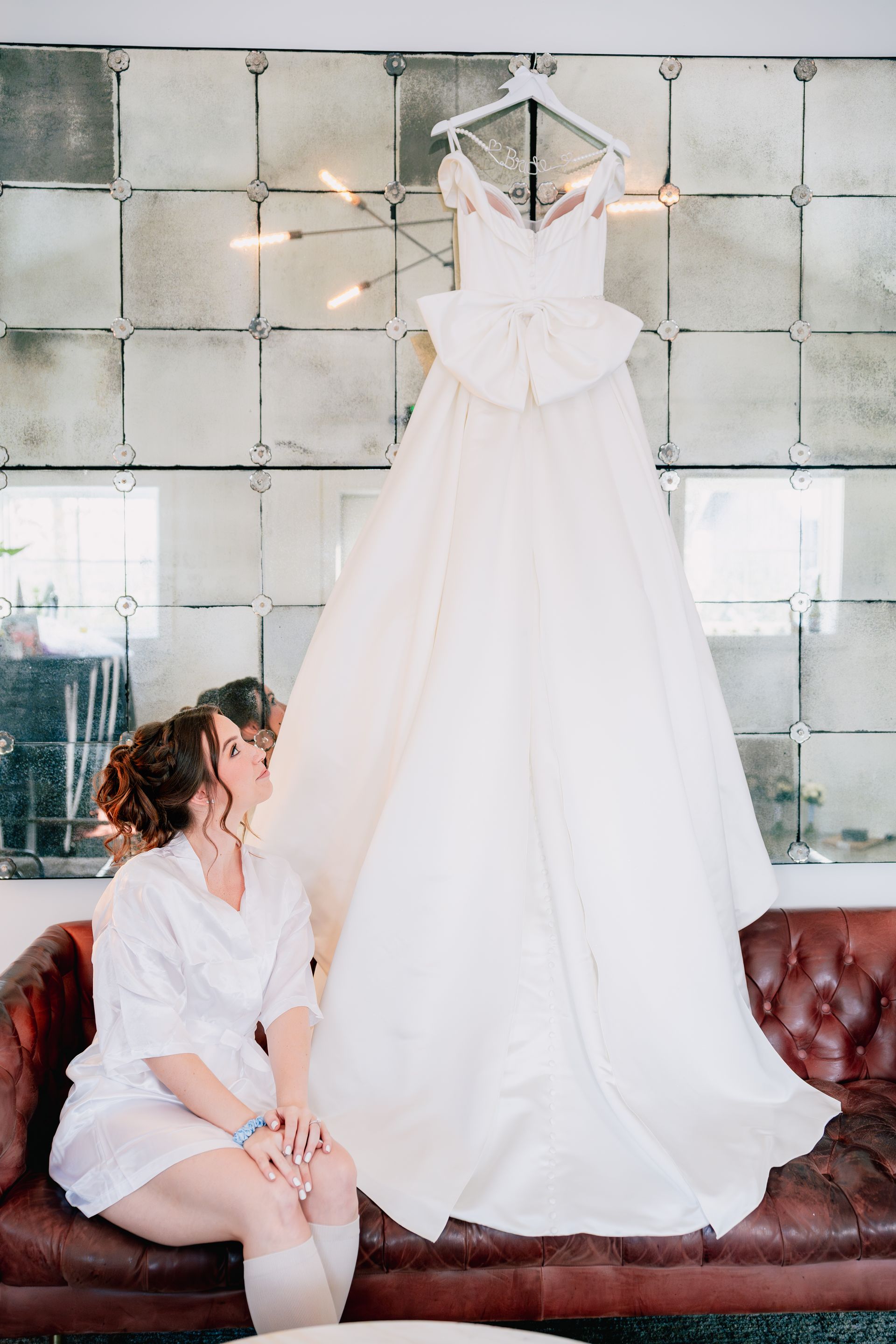 Wedding dress hanging on wall, bride sits on couch looking up at dress.