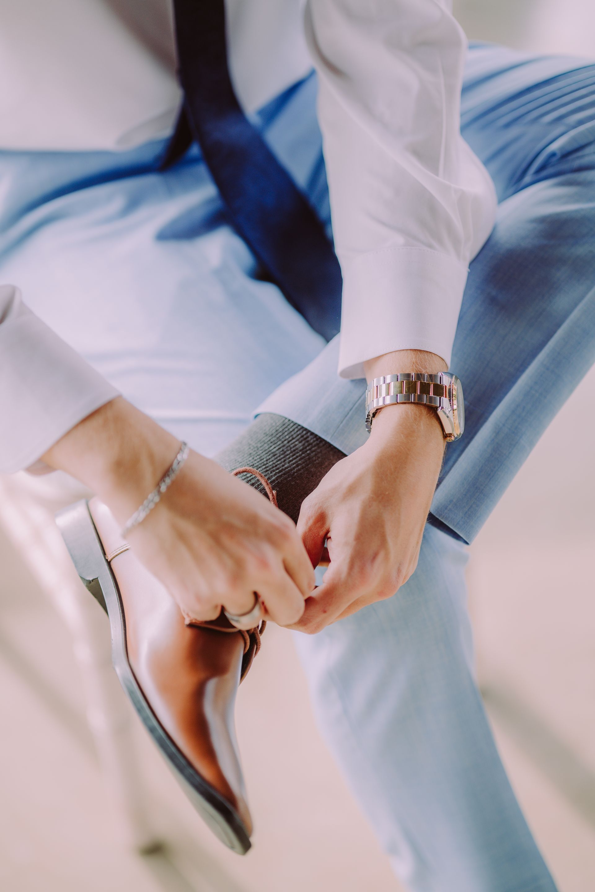 Man in light blue suit tying brown shoe; wrist watch and silver bracelet visible.