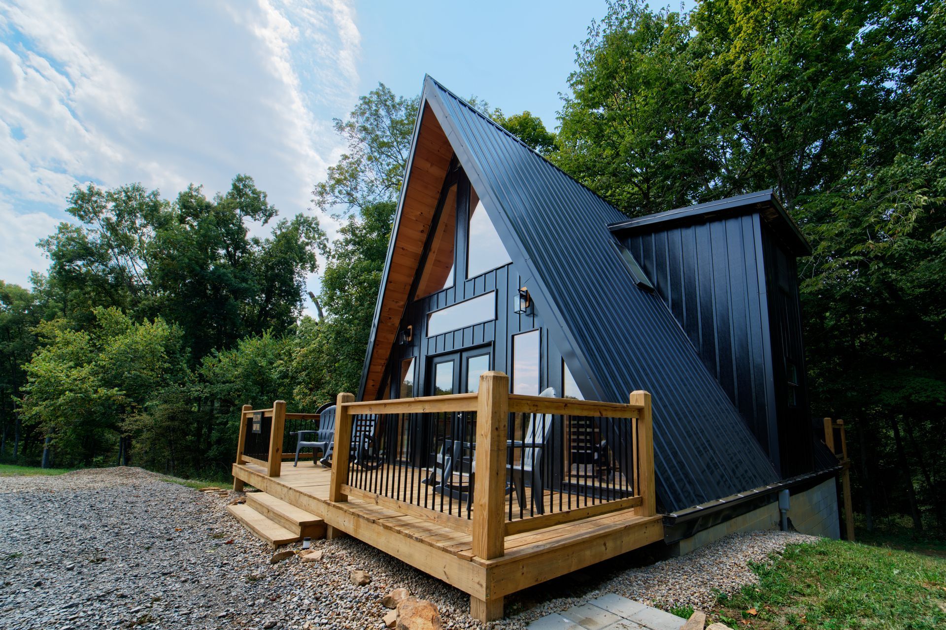 A black a frame cabin with a wooden deck in the woods.