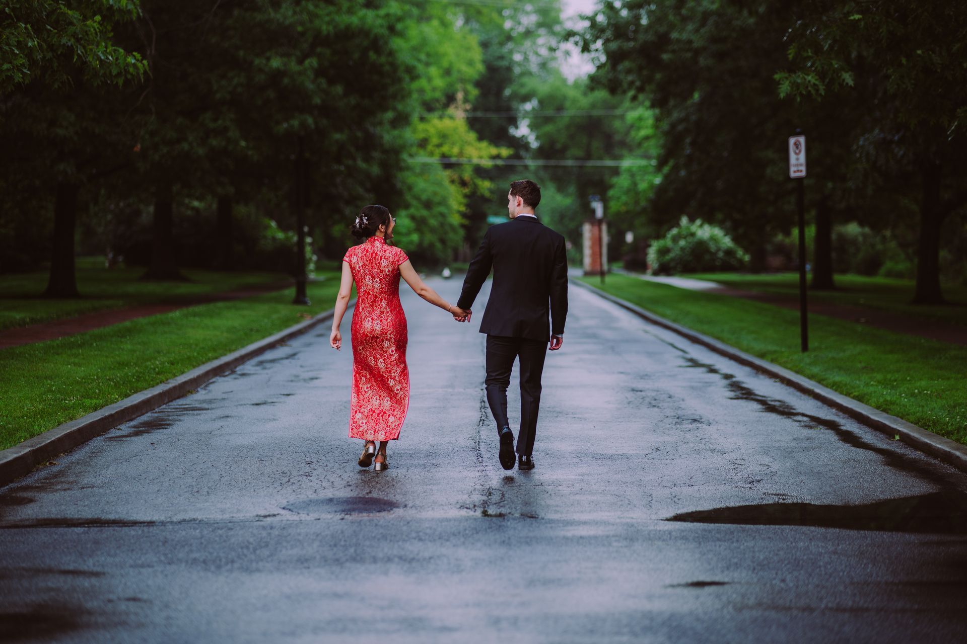 Two people holding hands and walking down a wet tree-lined road in the rain