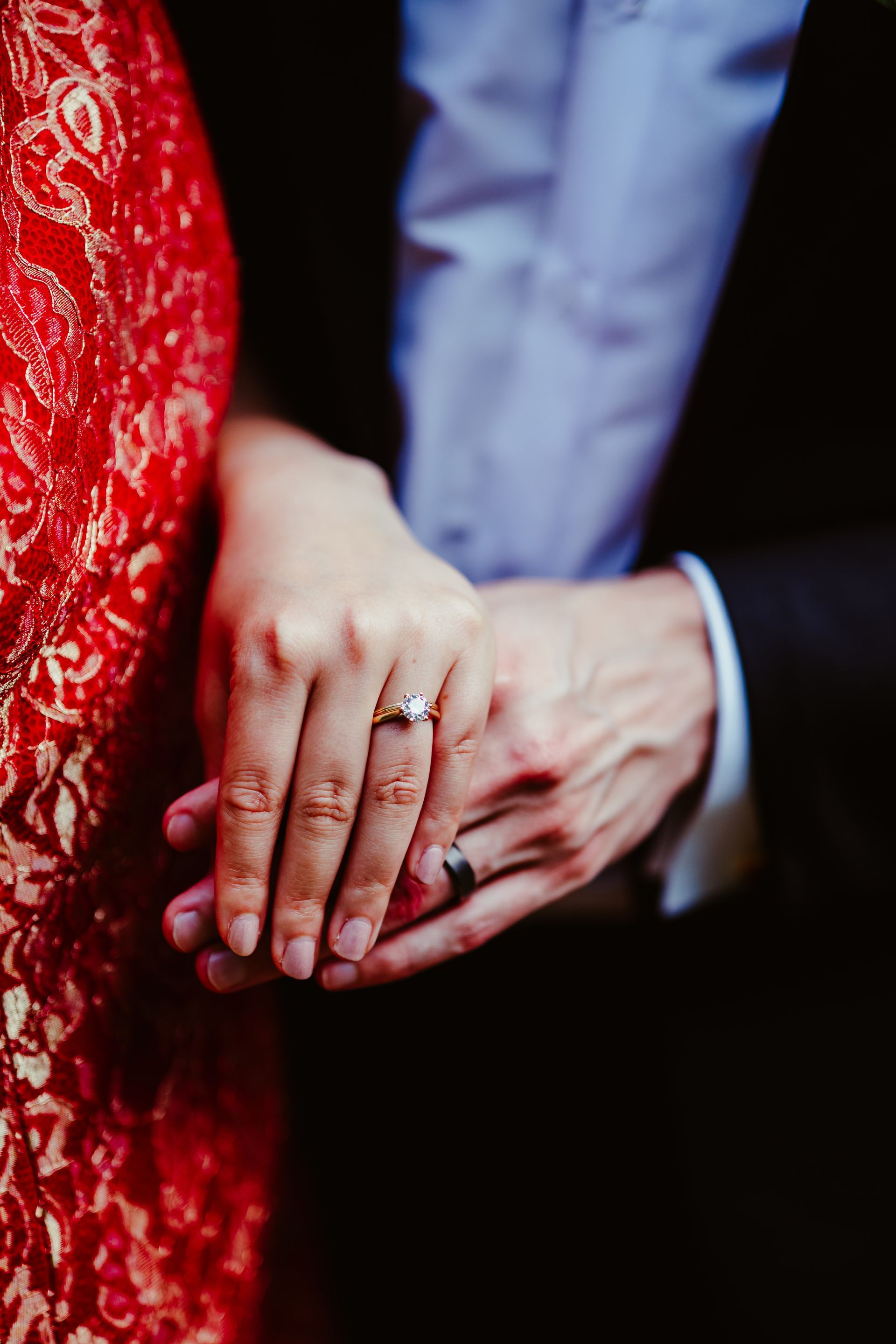 Hands clasped, one in a red lace dress and the other in a dark suit, with wedding rings visible.