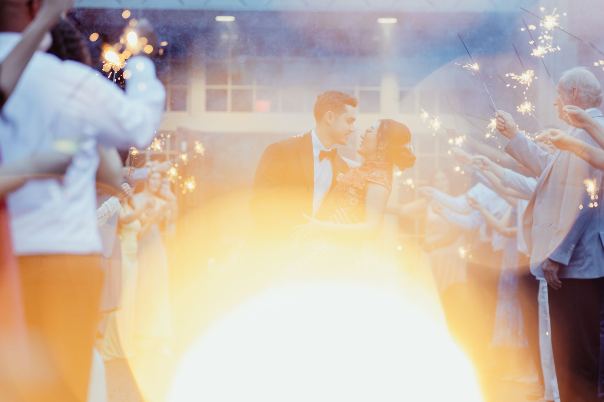 Couple sharing a wedding kiss amid sparklers and bright golden light at a celebration