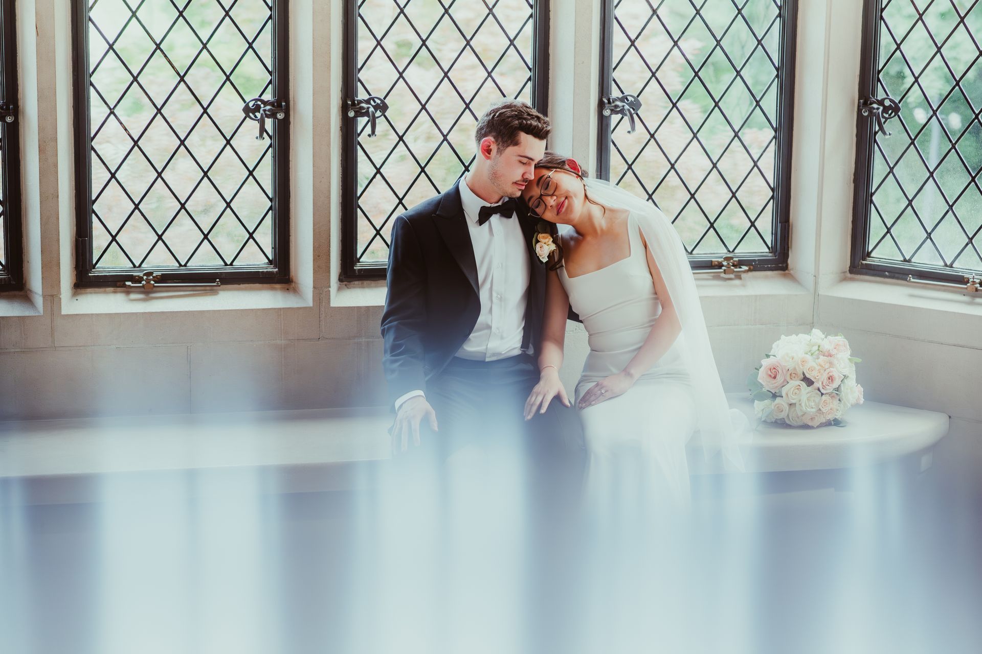 Bride and groom seated by tall windows, leaning together in a bright, airy room