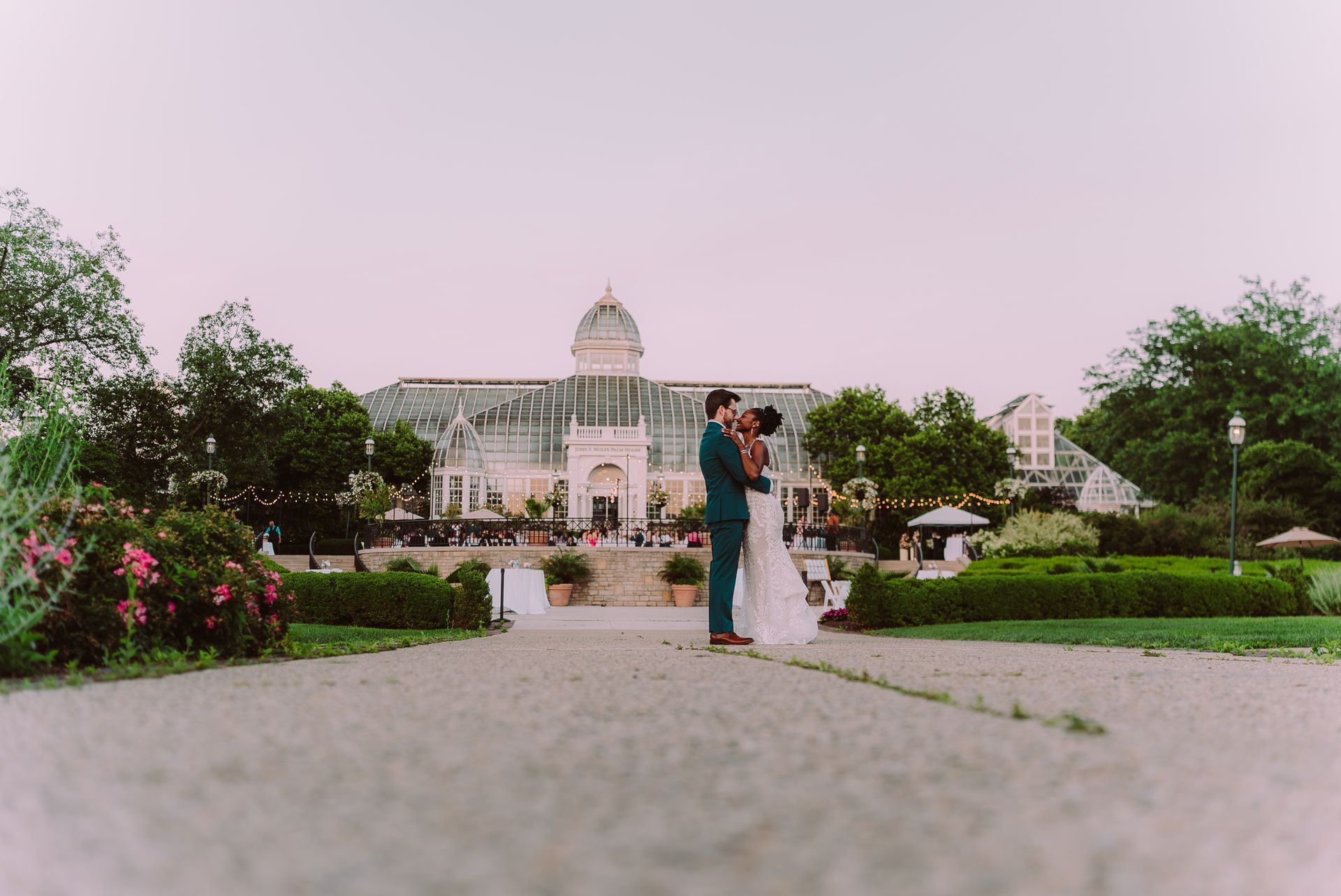 Bride and groom kissing in front of a glass greenhouse; sunset.