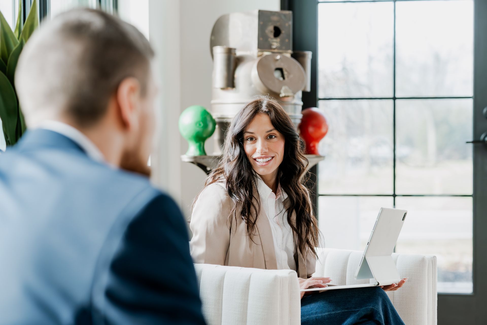 Woman with laptop smiles, conversing with a person in a blue suit in a bright room. | Photos by Kodjoarts