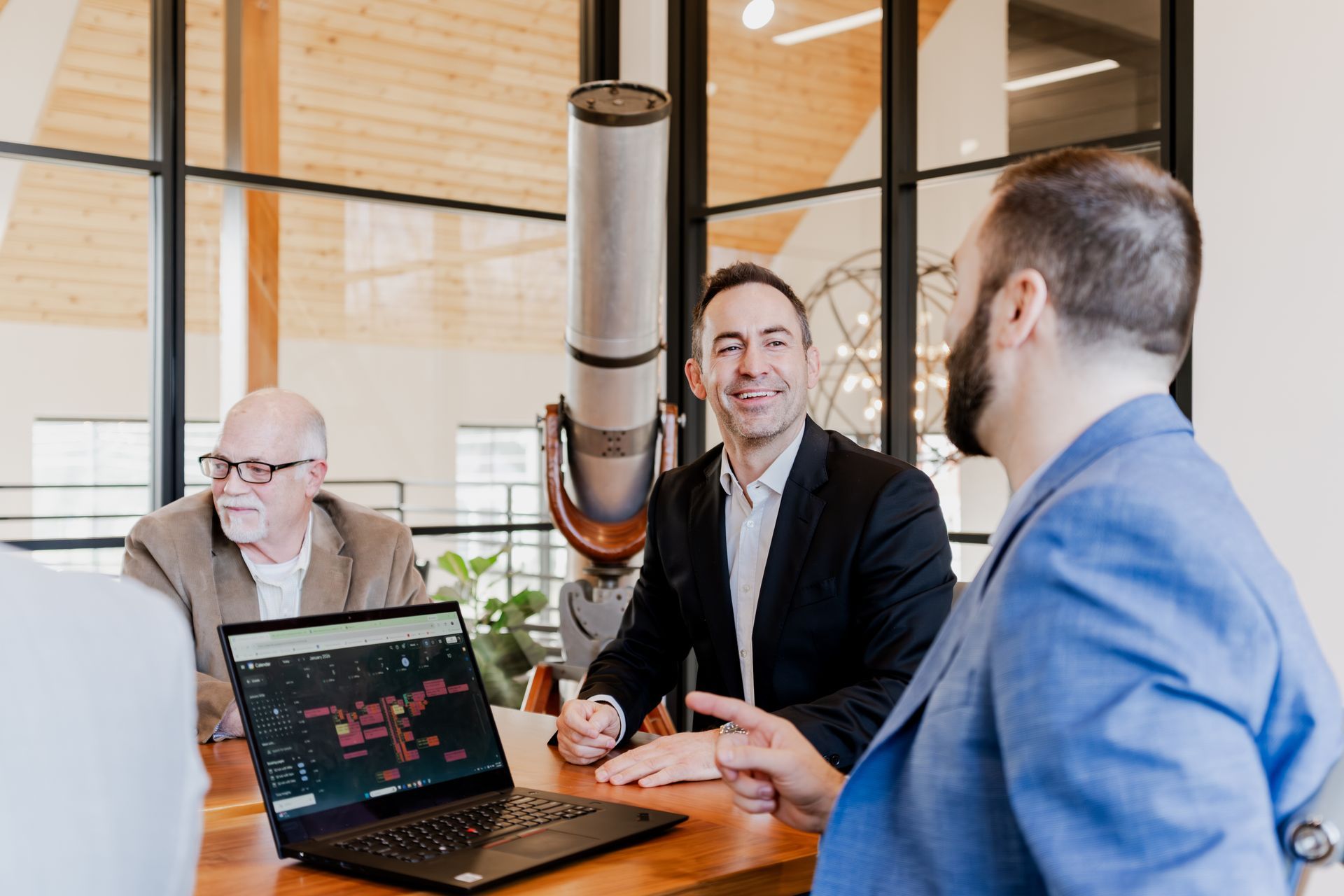 Business meeting around a table with laptop showing data. Men in suits, some smiling, in a modern office. | Photos by Kodjoarts