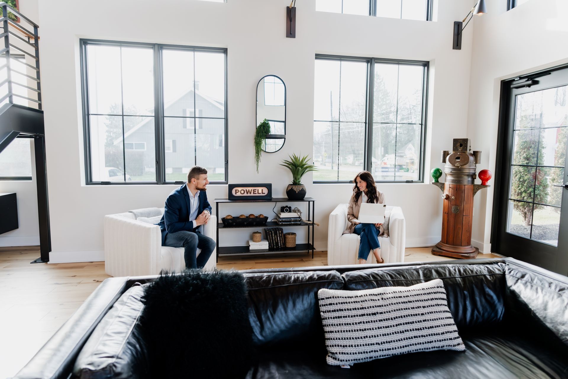 Two people seated, one using a laptop, in a bright, modern room with large windows.