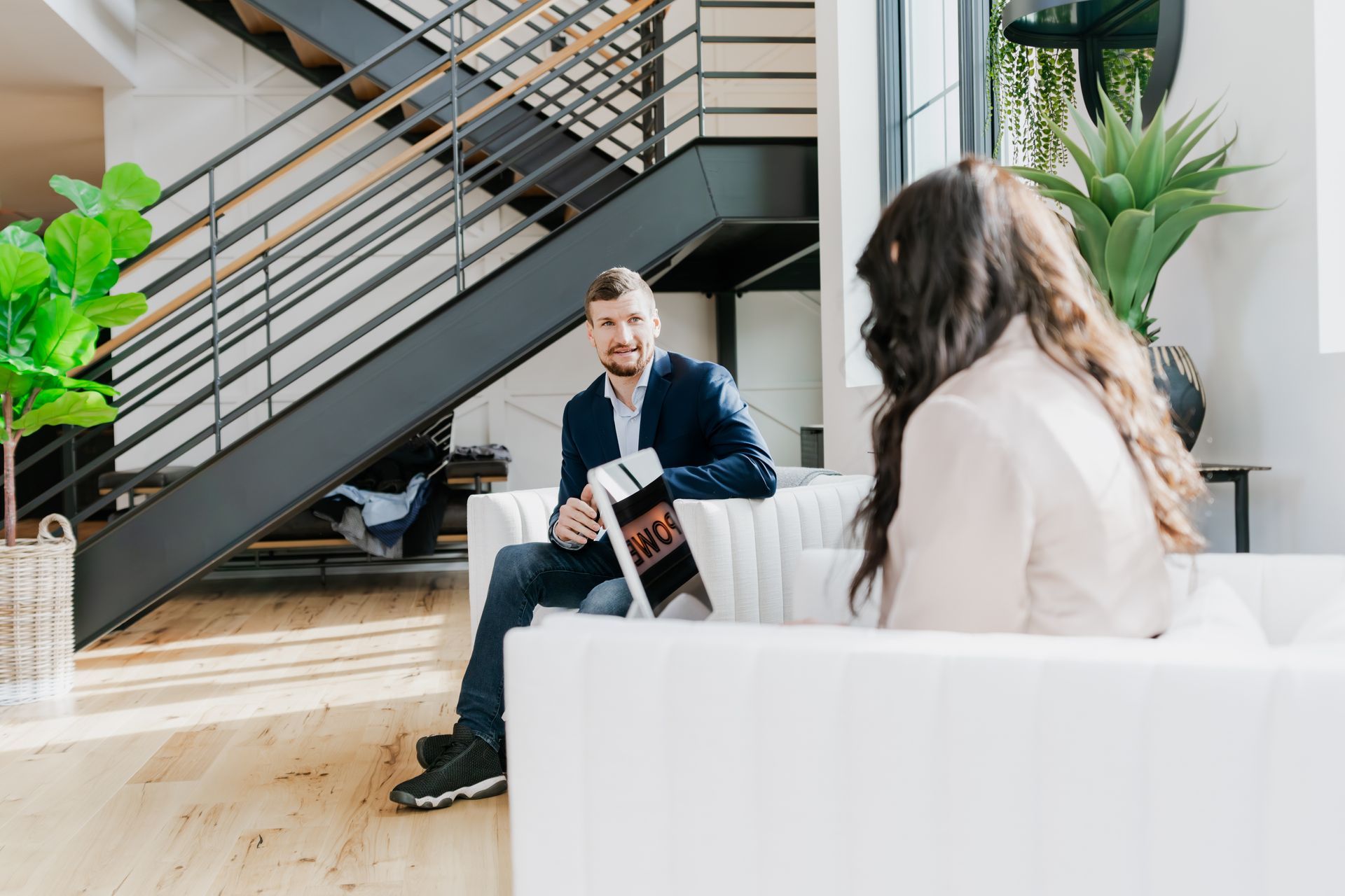 Man in blue blazer reviews documents with woman on white couch. Modern office interior. | Photos by Kodjoarts