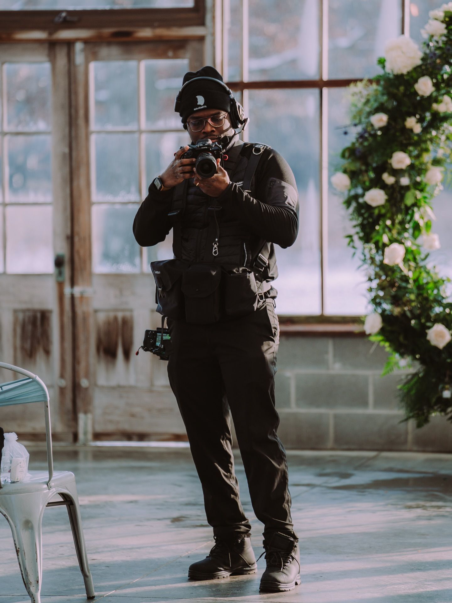 Man in black clothing and beanie films with a camera indoors, near a floral arch.