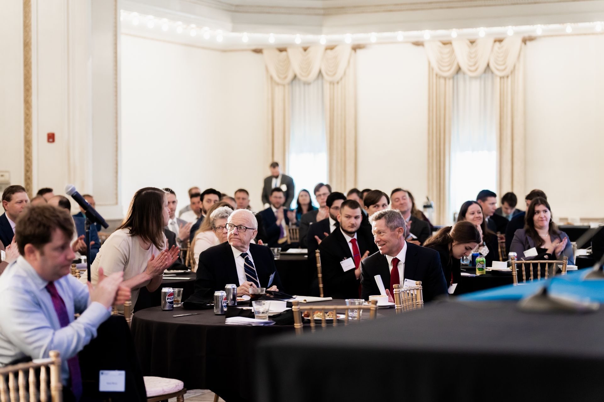 Audience members at round tables in a bright conference hall, some clapping, listening to a presentation.