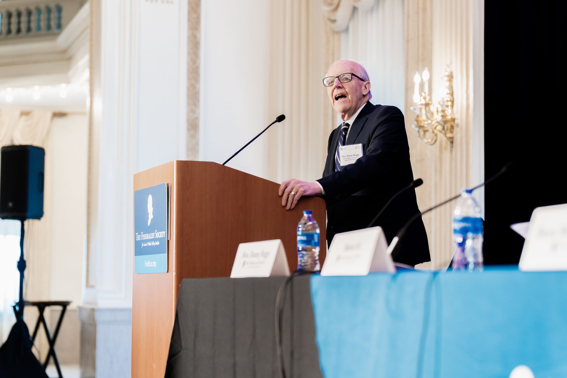 A person in a suit speaks from a podium at a formal indoor event.