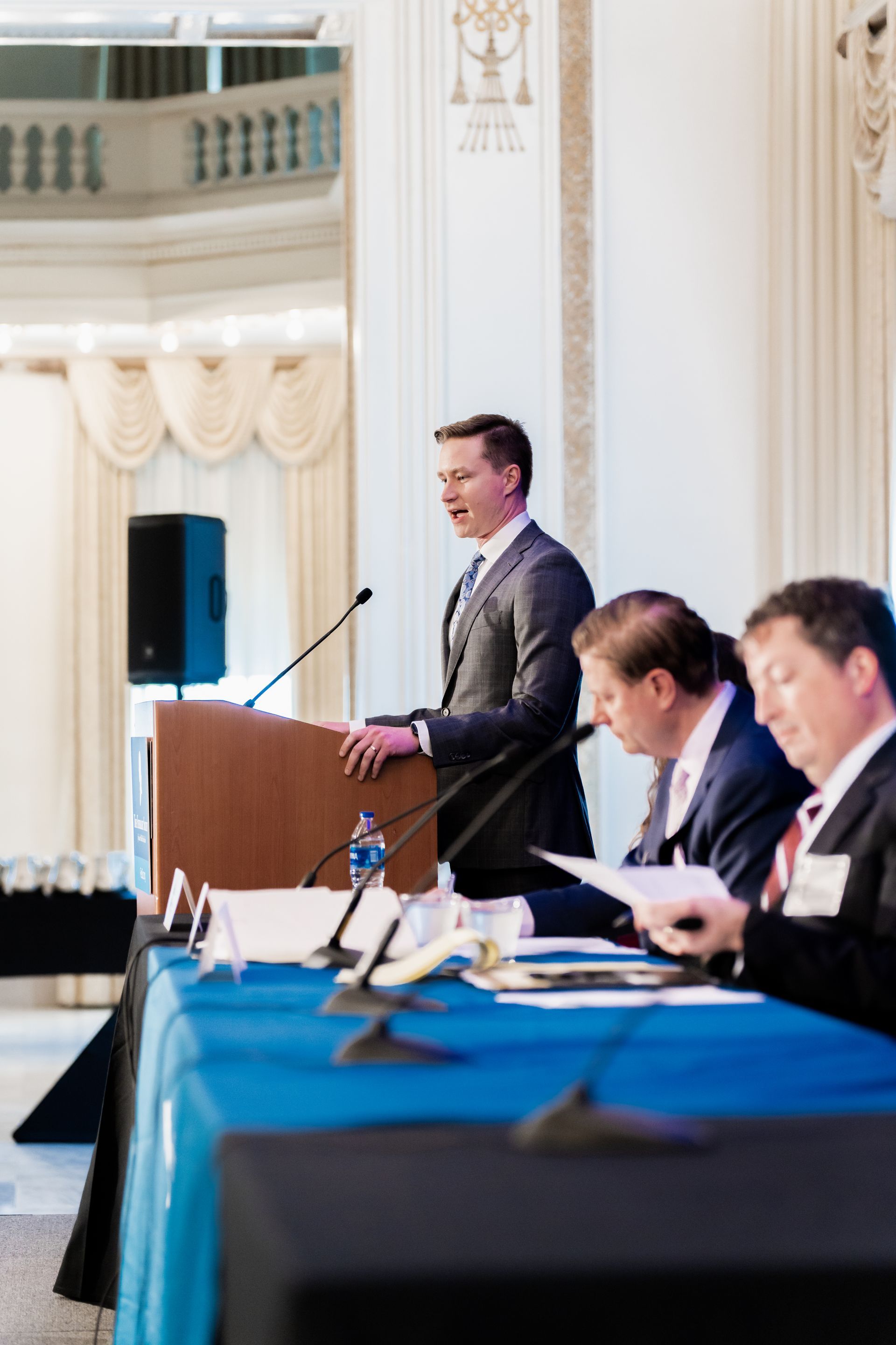 A speaker stands at a wooden podium before two men seated at a blue-covered table, set in a brightly lit indoor space.
