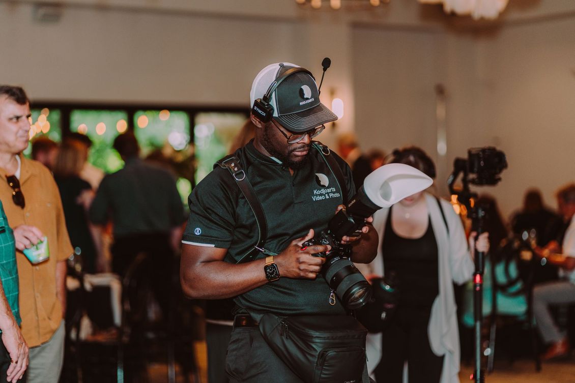 A photographer in a black uniform adjusts a camera indoors at an event, with other guests blurred in the background.