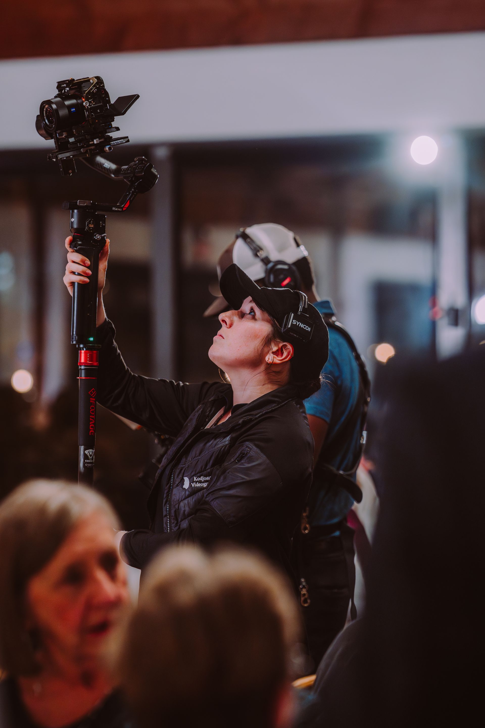 Woman operating a camera on a pole, indoors, looking up. Another person stands behind her.