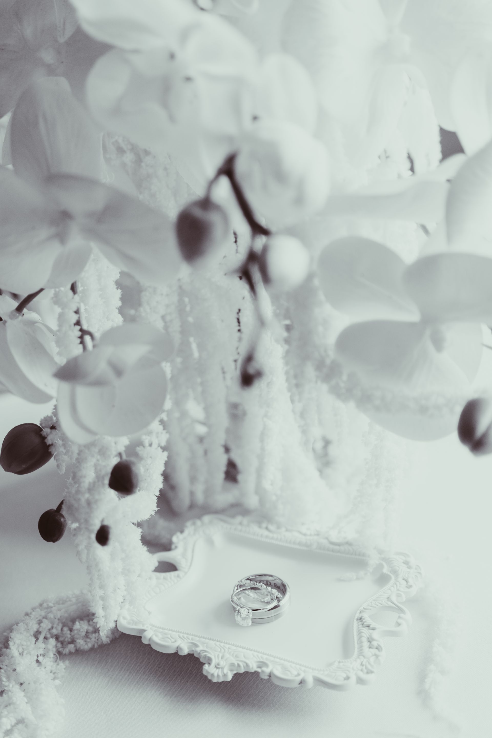 A black and white photo of a wedding ring on a pillow surrounded by flowers.