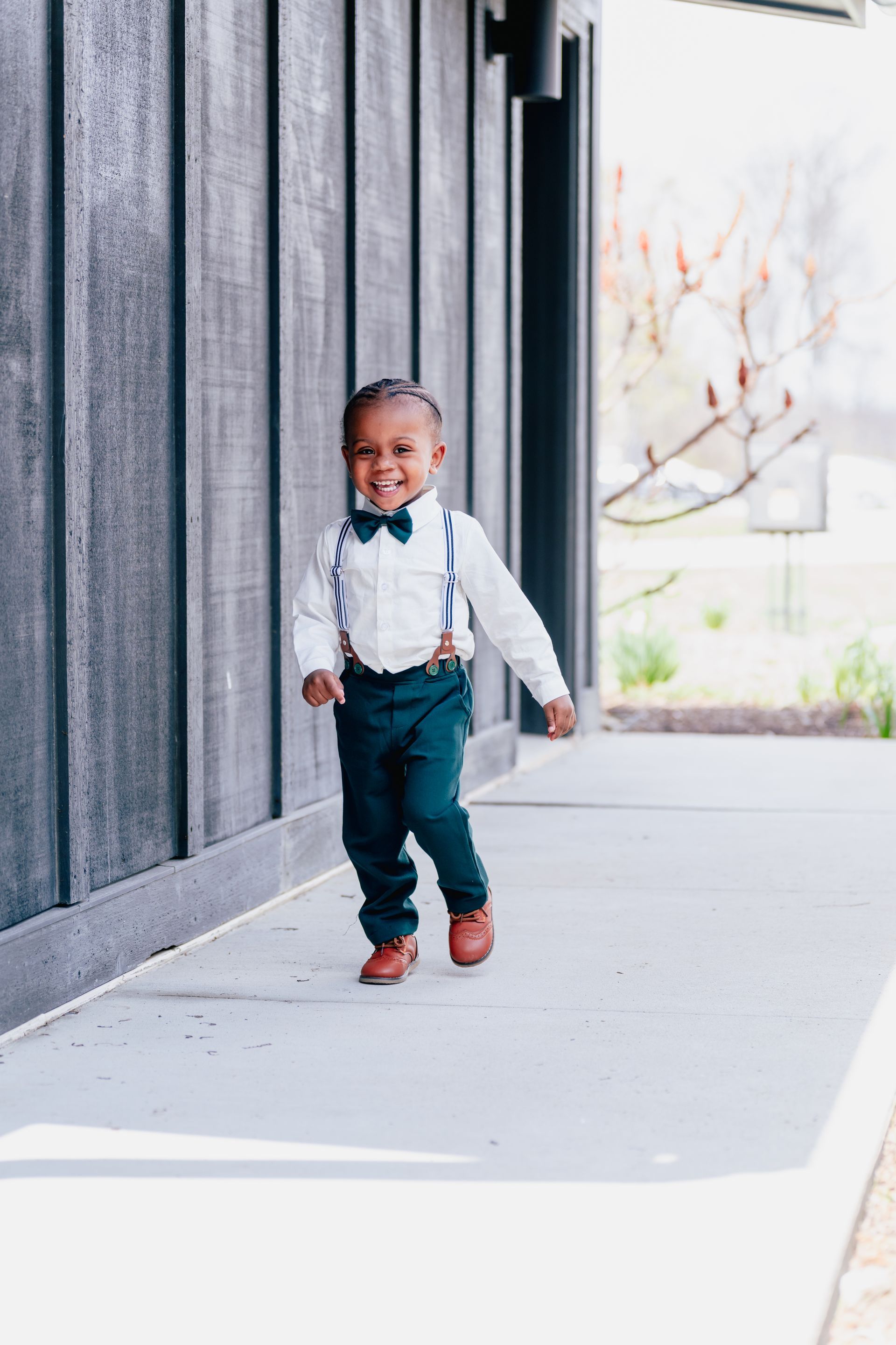 A little boy wearing suspenders and a bow tie is walking down a sidewalk.