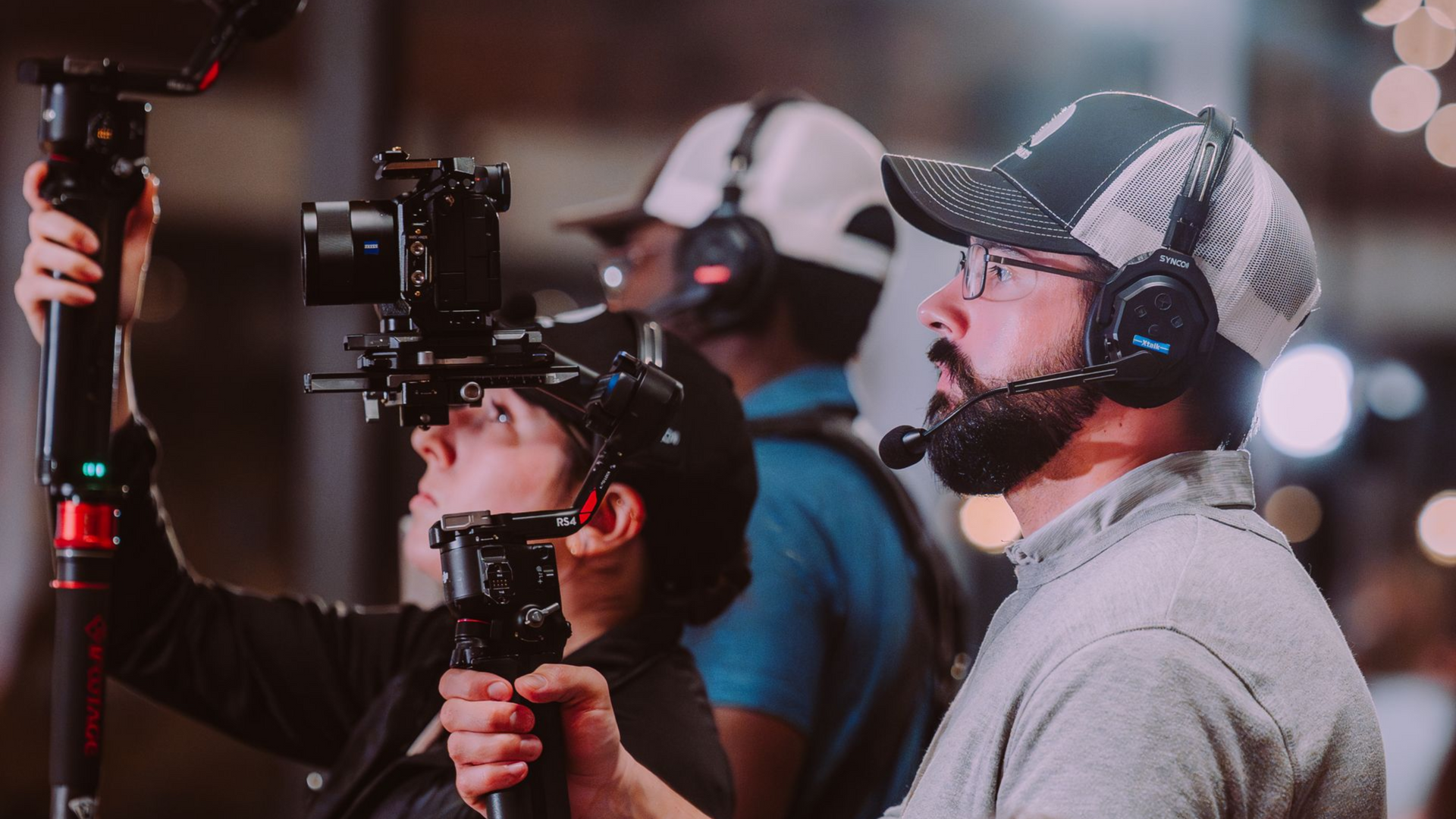 Three people wearing headsets, operating video cameras, indoors with bokeh background.