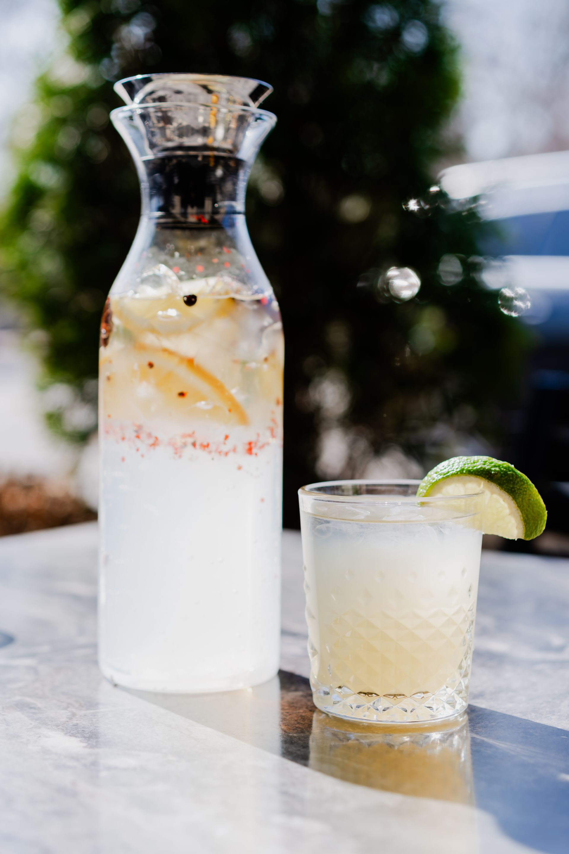 Cocktail and pitcher on a marble surface with a blurred green tree background. The cocktail is garnished with a lime.