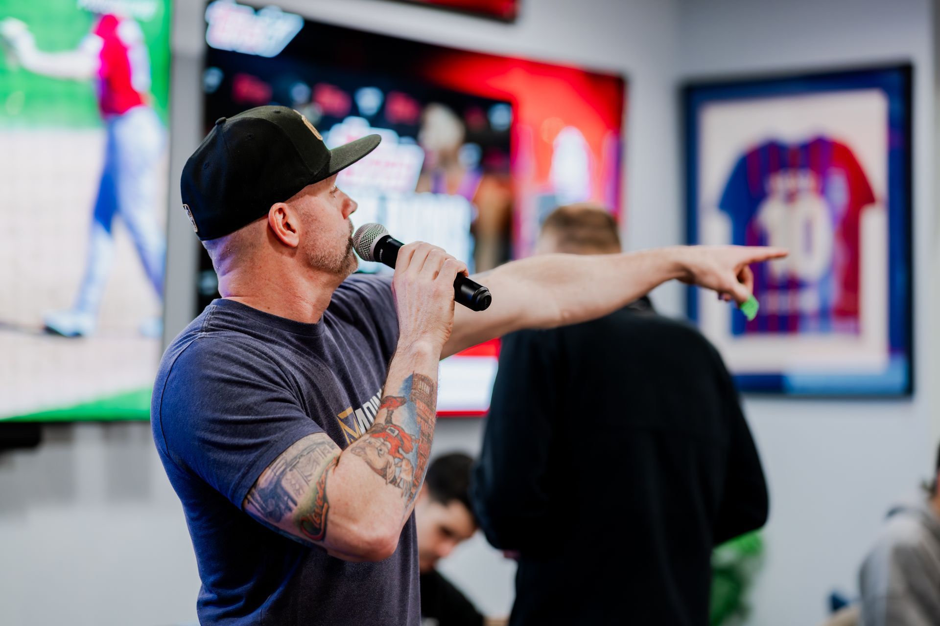 Man in a black cap with a microphone gestures right in front of a group of people. Midwest Card | Event Photography