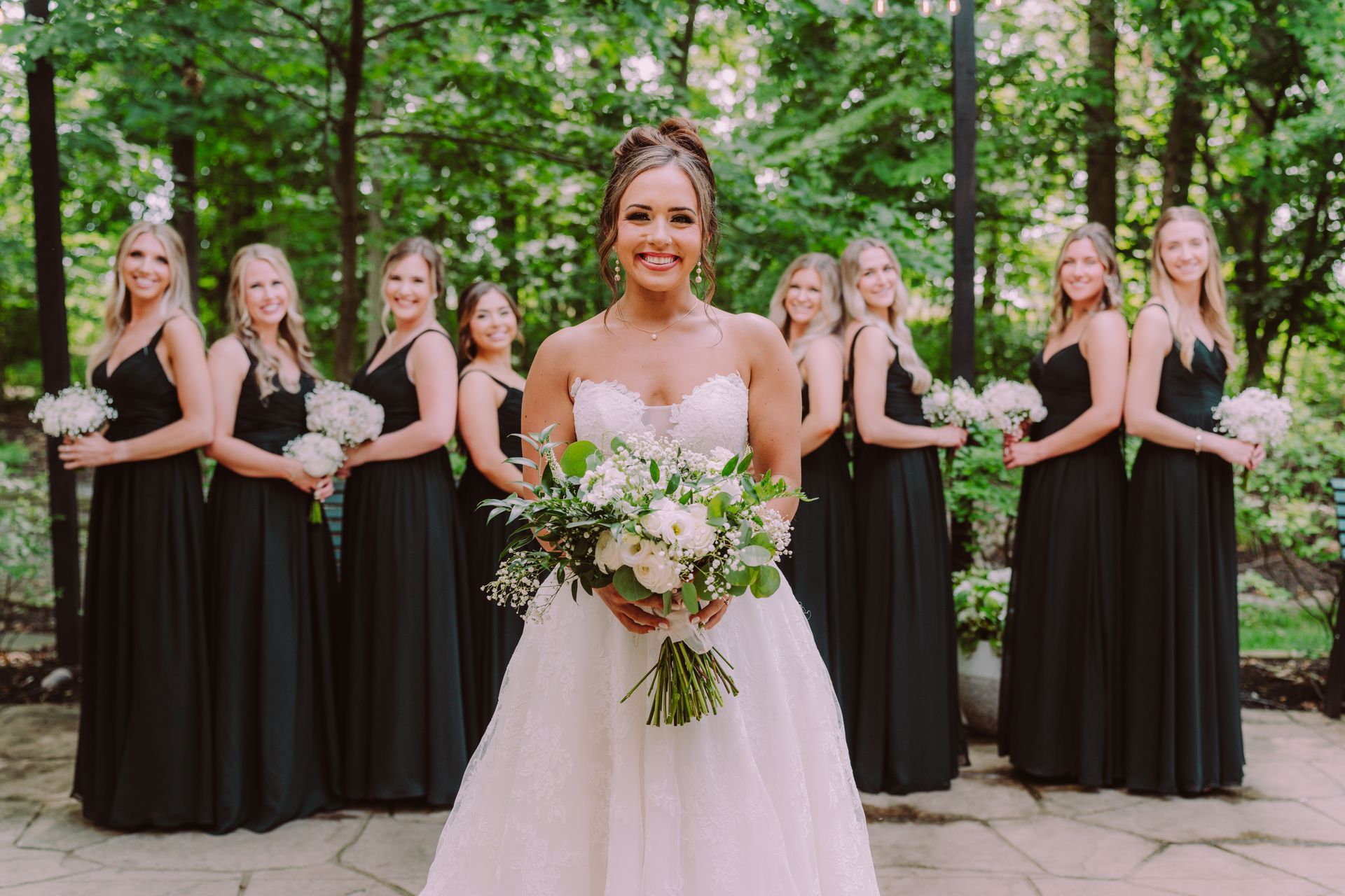Bride in a white gown, holding a bouquet, smiles with her bridesmaids in black dresses. They stand in front of a wooded backdrop.