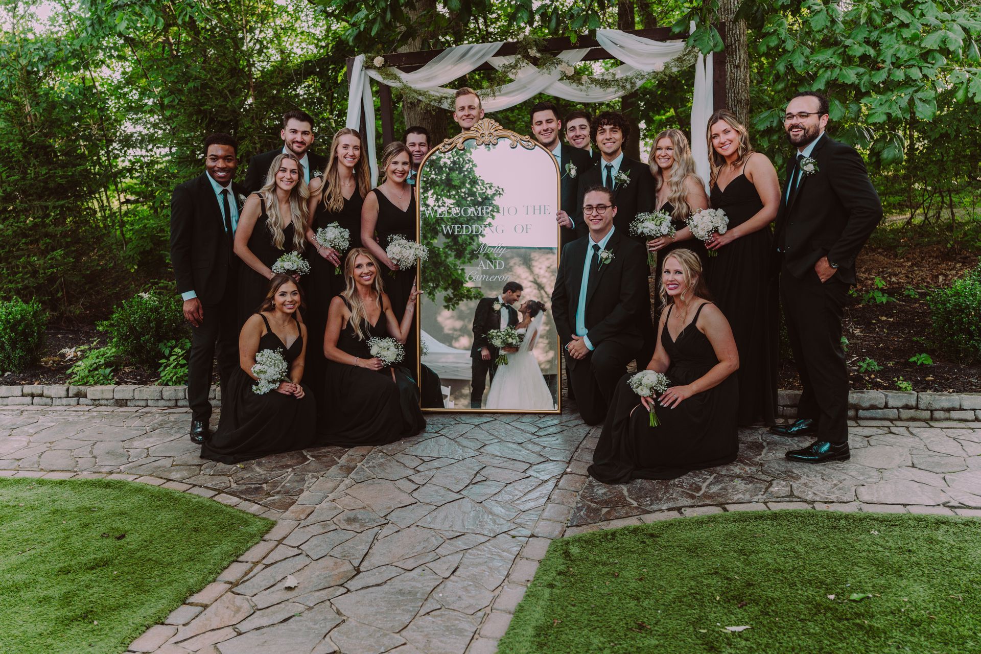 Wedding party posing with a mirror reflecting the bride and groom. People in black dresses and suits stand and kneel on a stone path, outdoors.