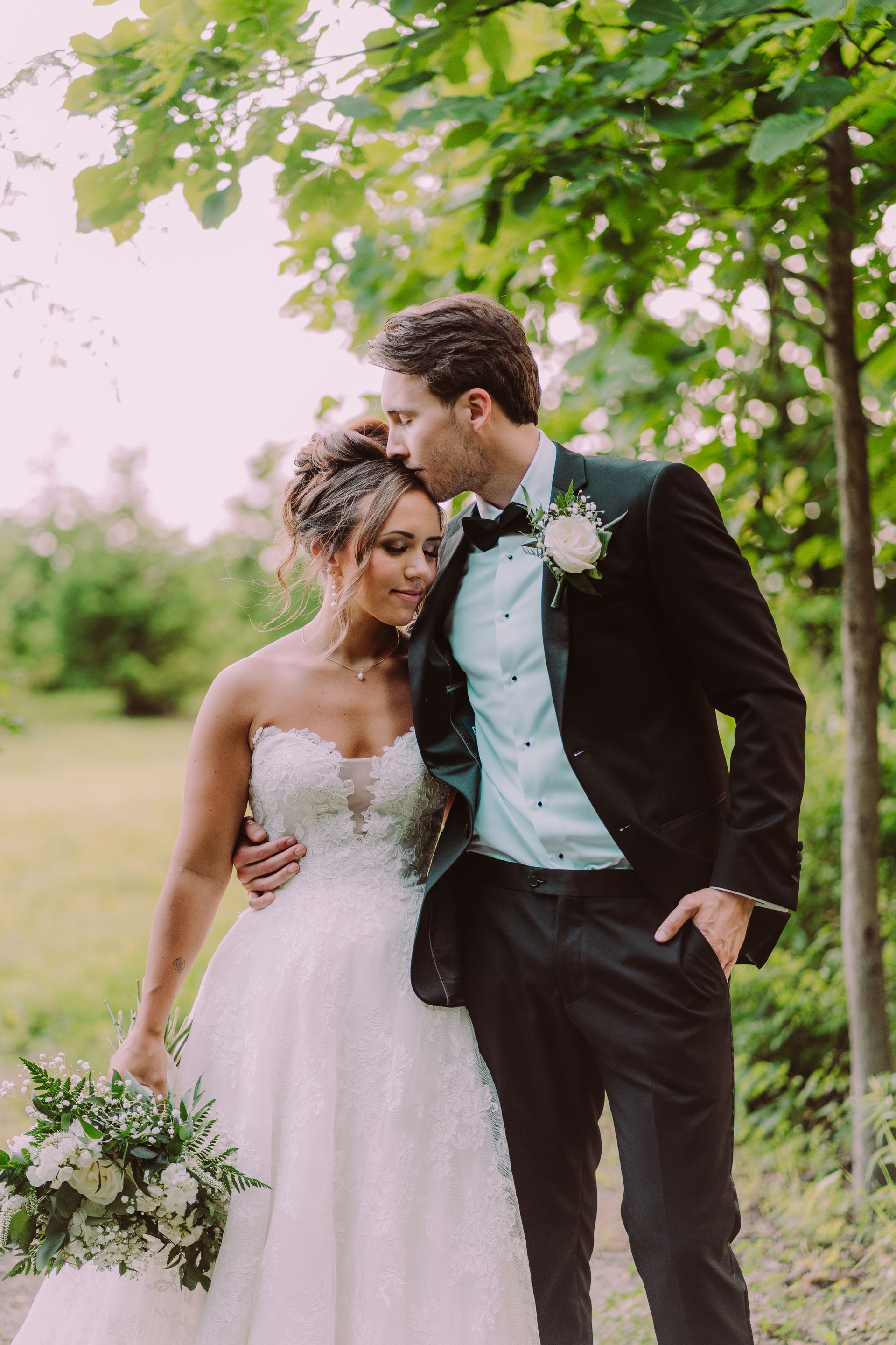 Bride and groom share a tender moment in a natural setting. The groom kisses the bride's forehead; she smiles while holding her bouquet.