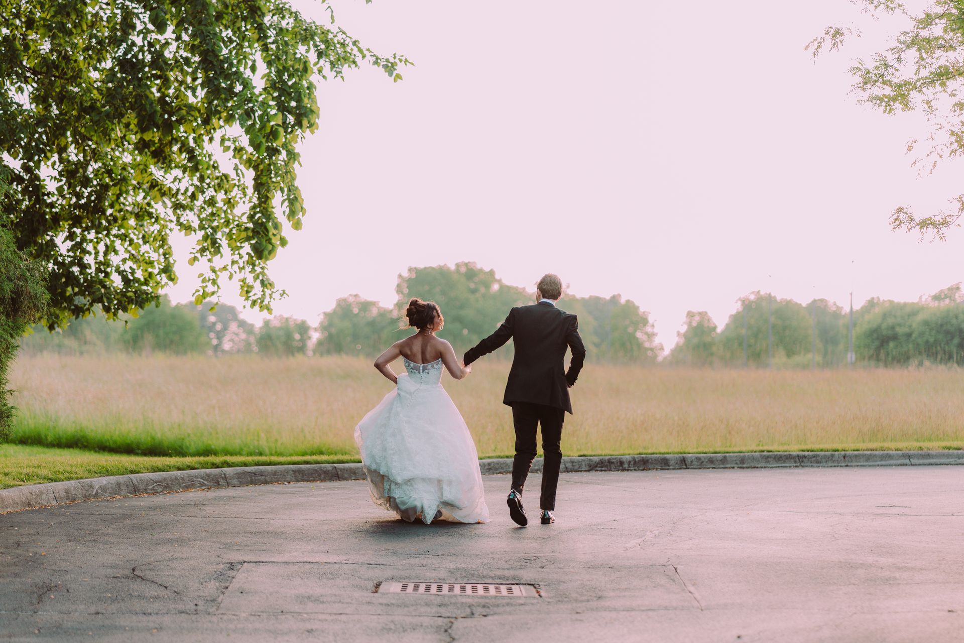 Newlyweds run hand-in-hand across a paved area towards a field. The bride wears a white gown; the groom, a black suit. Soft sunlight bathes the scene.