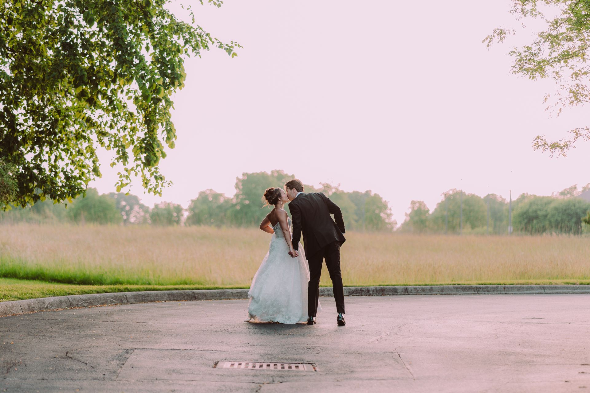 Newlyweds share a kiss outdoors. The bride wears a white gown, and the groom is in a black suit, standing in a paved area with a field in the background.