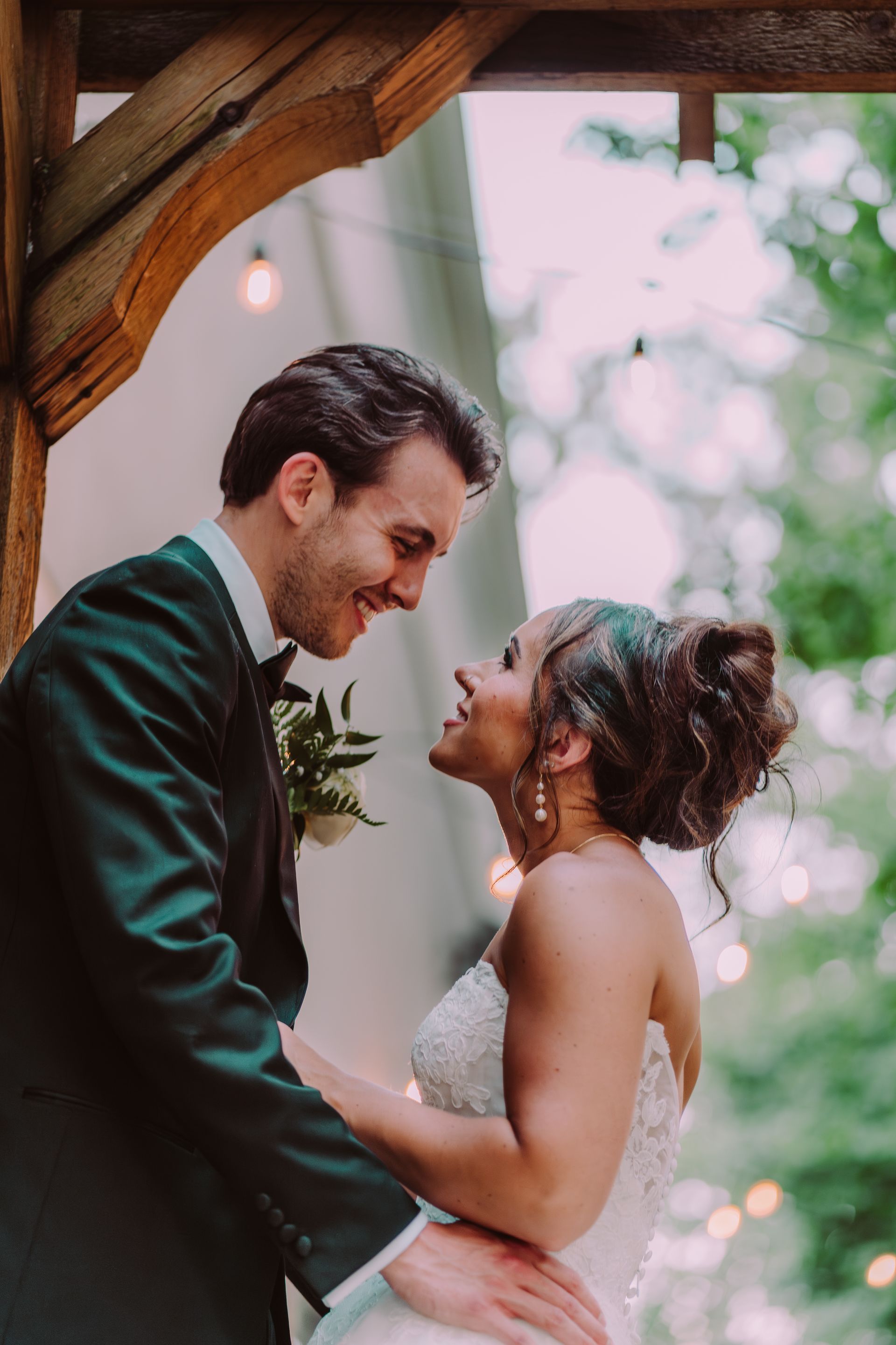 Bride and groom smiling at each other, standing under a wooden archway, outdoor setting. Man in a black tuxedo, woman in a white wedding dress.