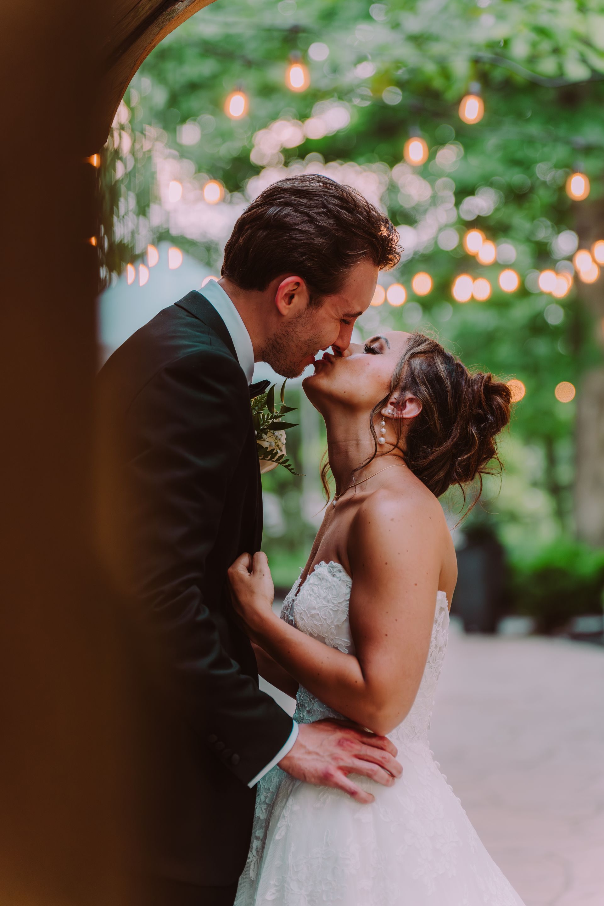 Bride and groom kissing under an archway decorated with lights. The bride wears a white strapless gown; the groom is in a black suit.