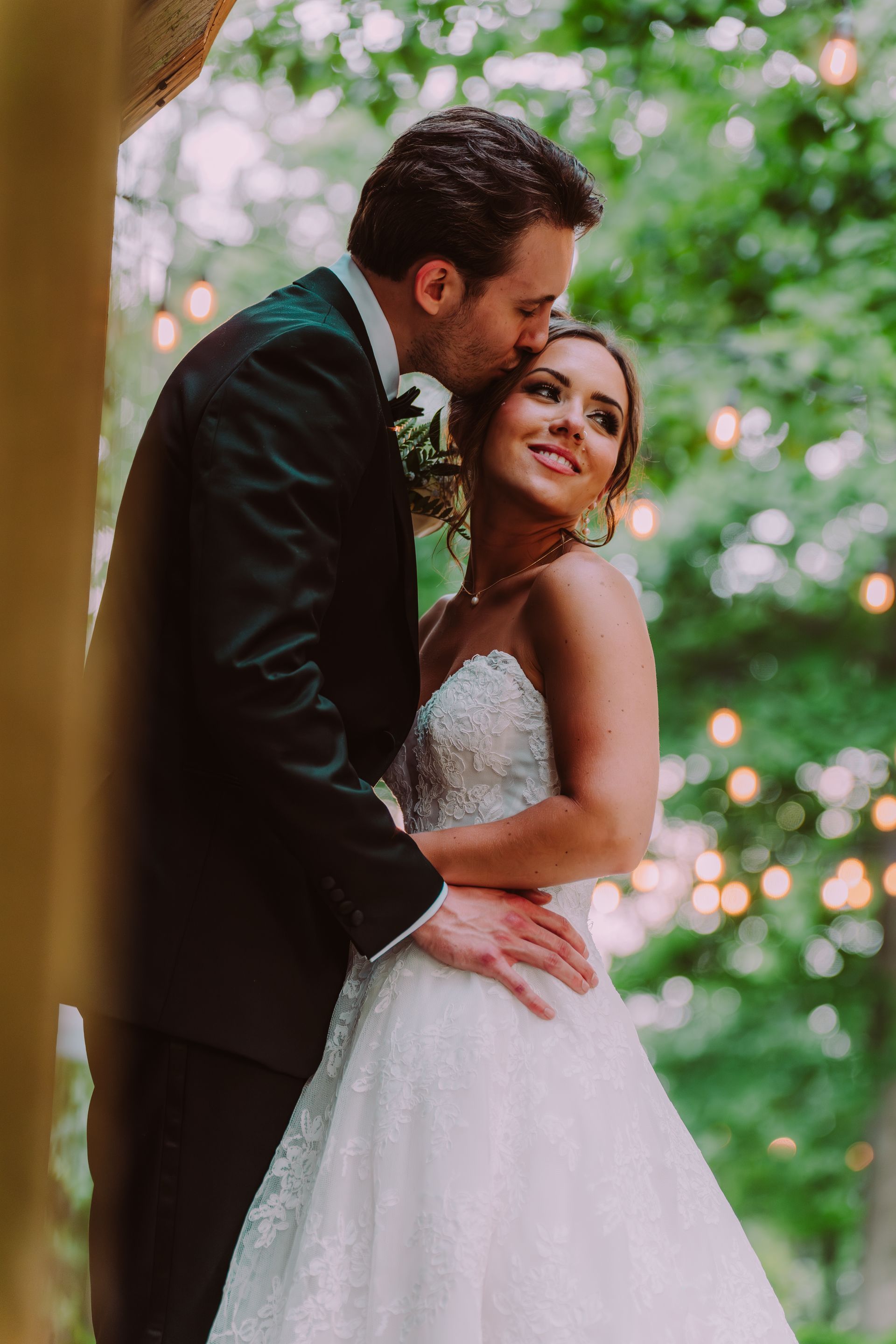 A groom in a black tuxedo kisses the forehead of his bride, who wears a white beaded gown, outside among trees with soft background lights.