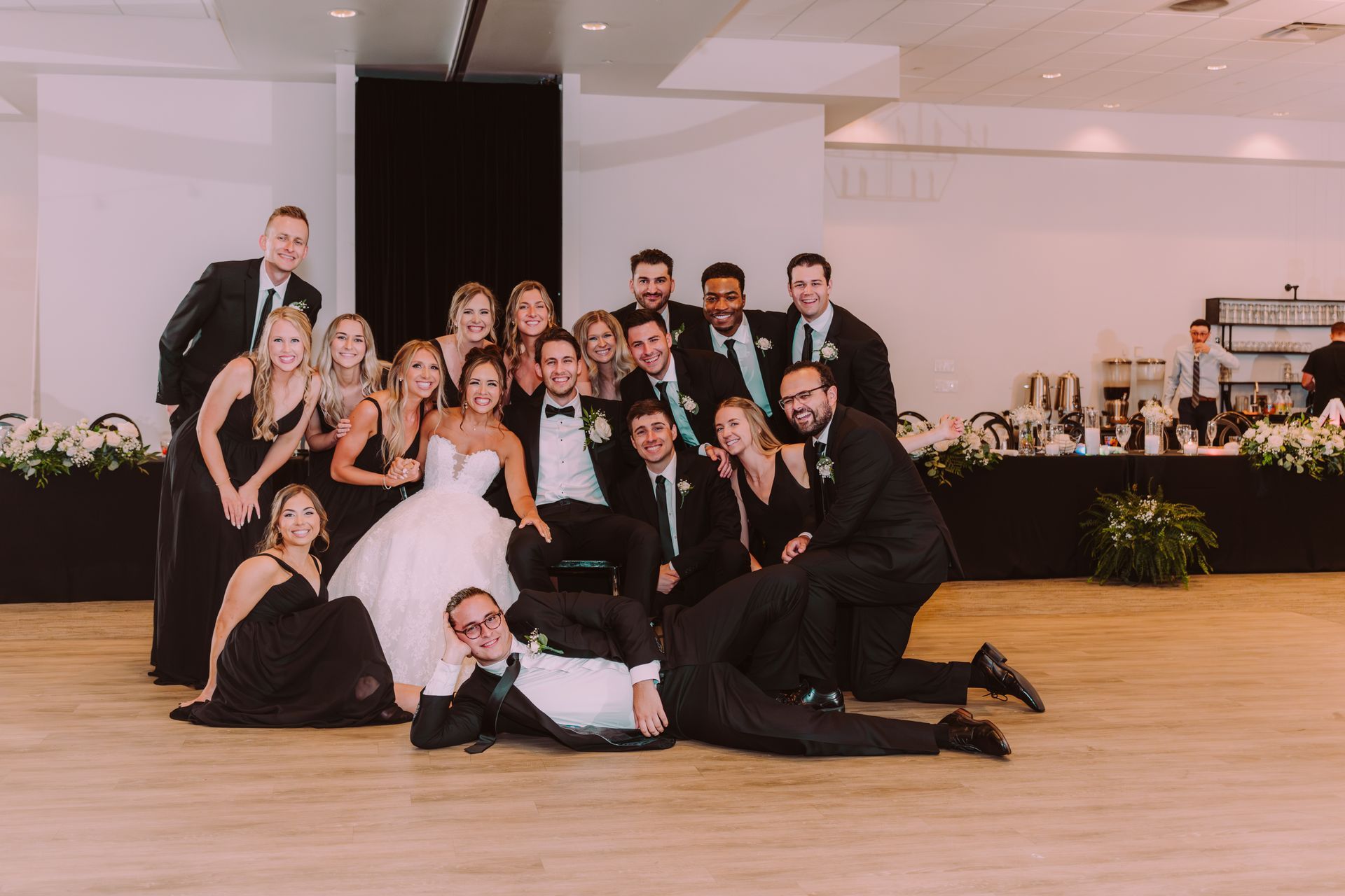 Wedding party group photo. Bride in white dress with bridesmaids in black. Groomsmen in black suits and ties. Posed indoors in a ballroom.