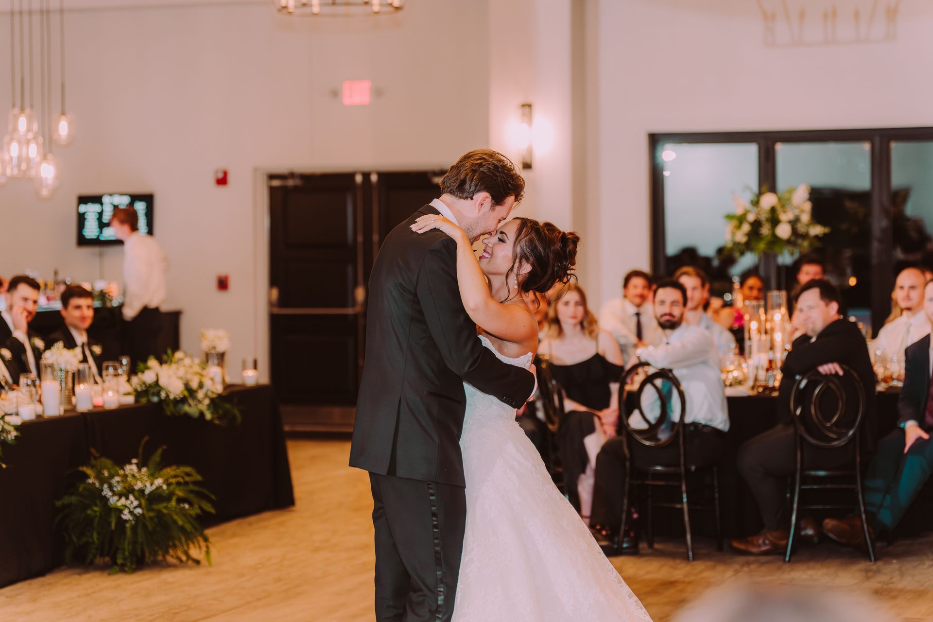 Newlyweds share a kiss while dancing at their wedding reception. The bride wears a white gown, and the groom is in a black suit. Guests watch in the background.