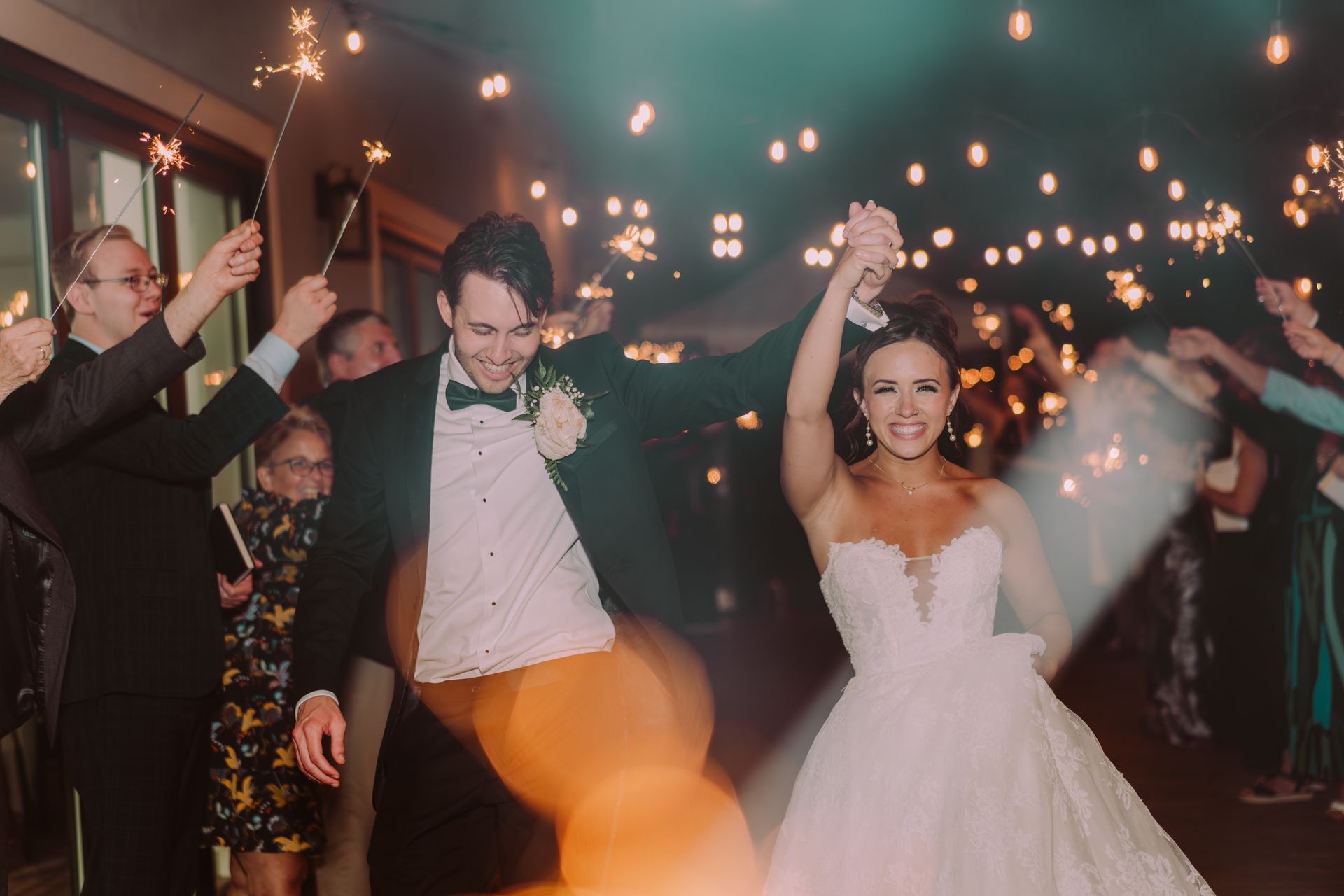 Newlyweds exit, holding hands, through a sparkler-lit tunnel, smiling. Nighttime, outdoor setting with guests cheering.
