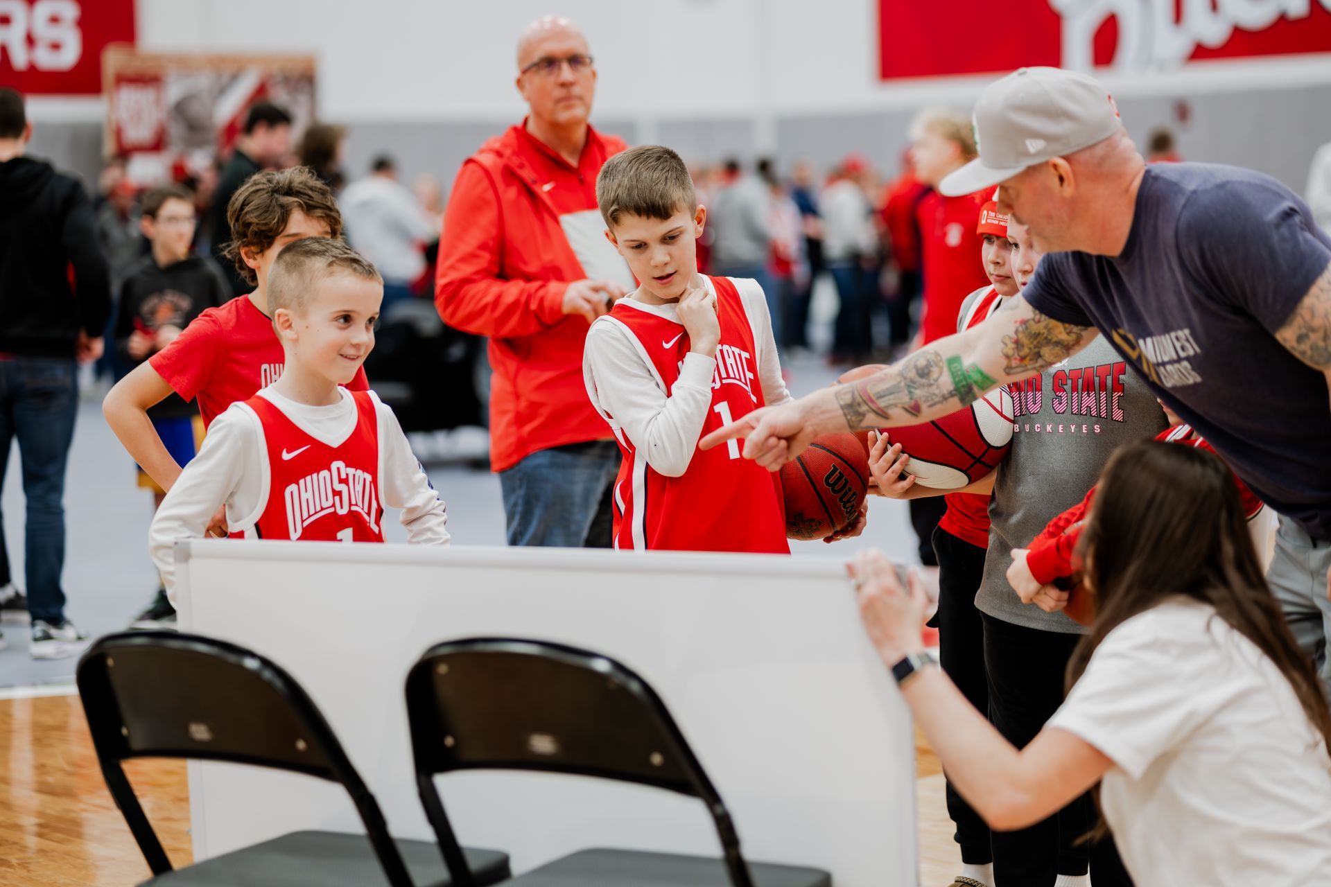 Boys in red jerseys with a basketball, interacting with adults near a white board.Midwest Cards | Event Photography