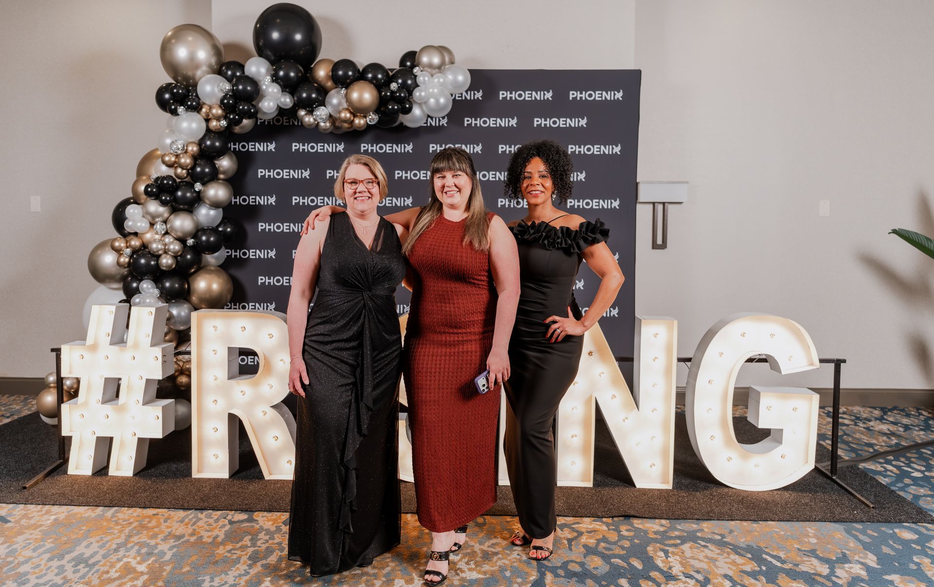 Three people pose smiling in front of a Phoenix-branded backdrop and large illuminated "RISING" letters.